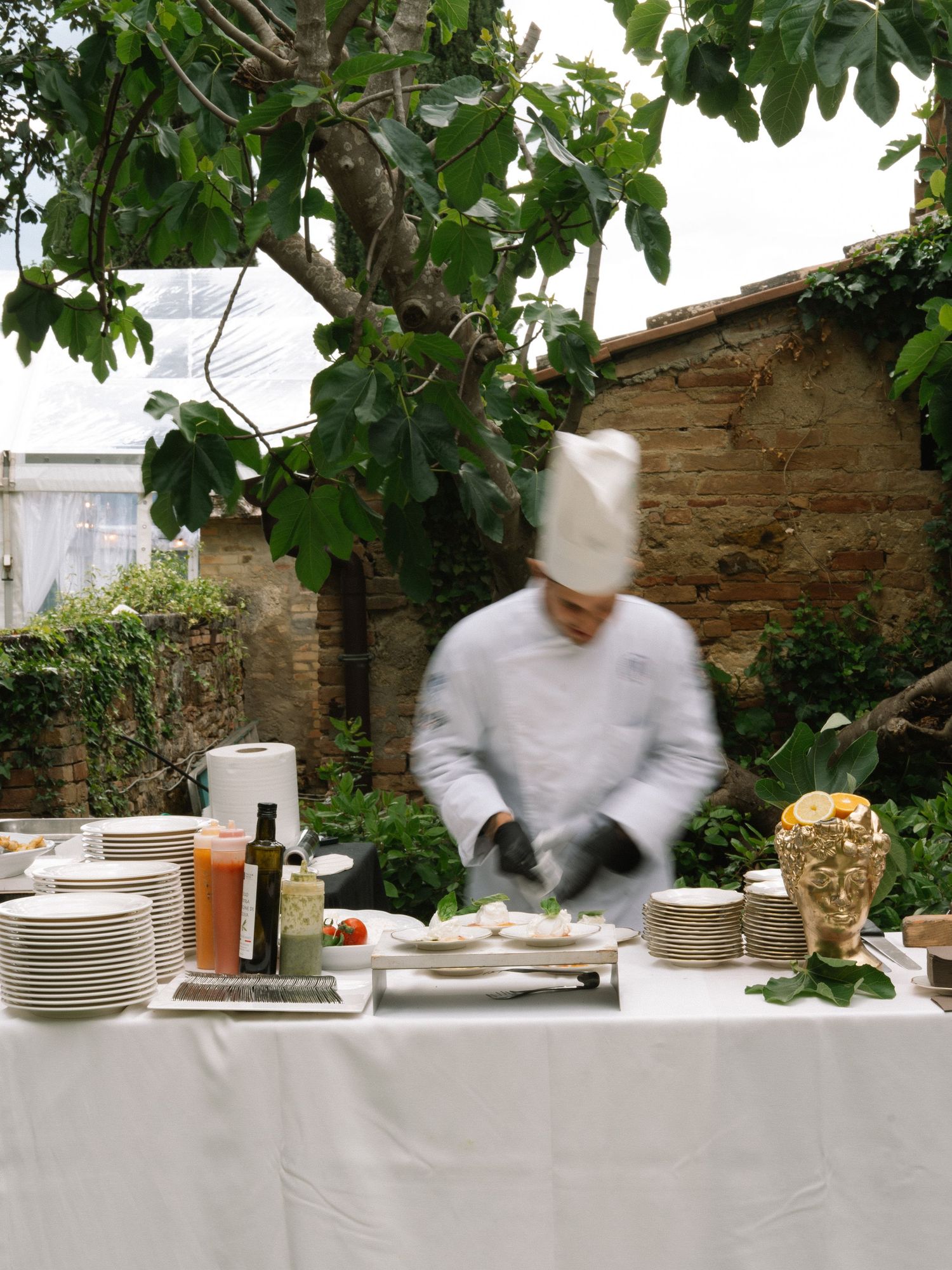Chef in white uniform and toque prepares dishes at an outdoor wedding catering station under a tree.