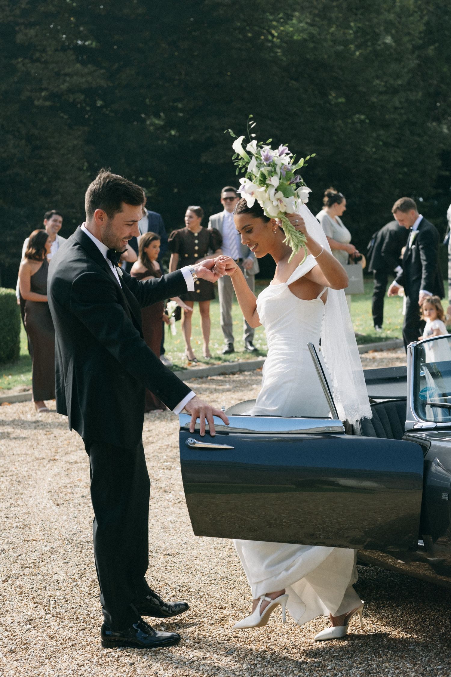 Groom helps bride out of vintage convertible car at outdoor wedding ceremony with guests in background.