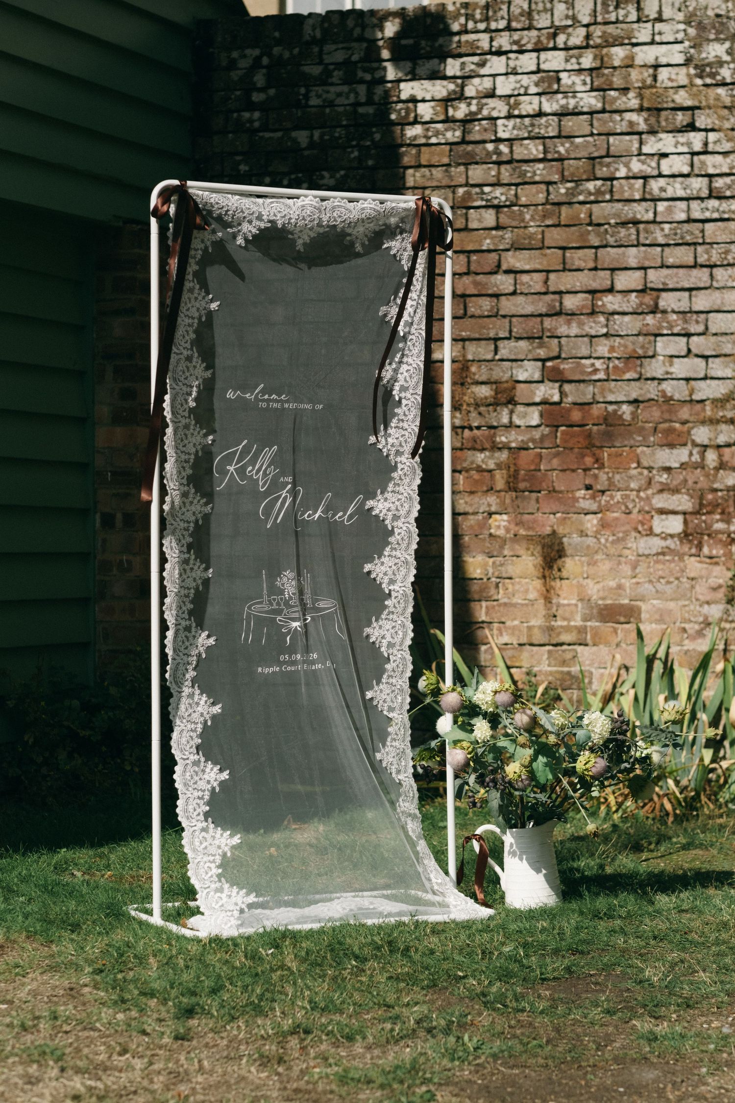 Elegant wedding welcome sign on sheer fabric with lace trim, displayed on a copper stand beside a floral arrangement.