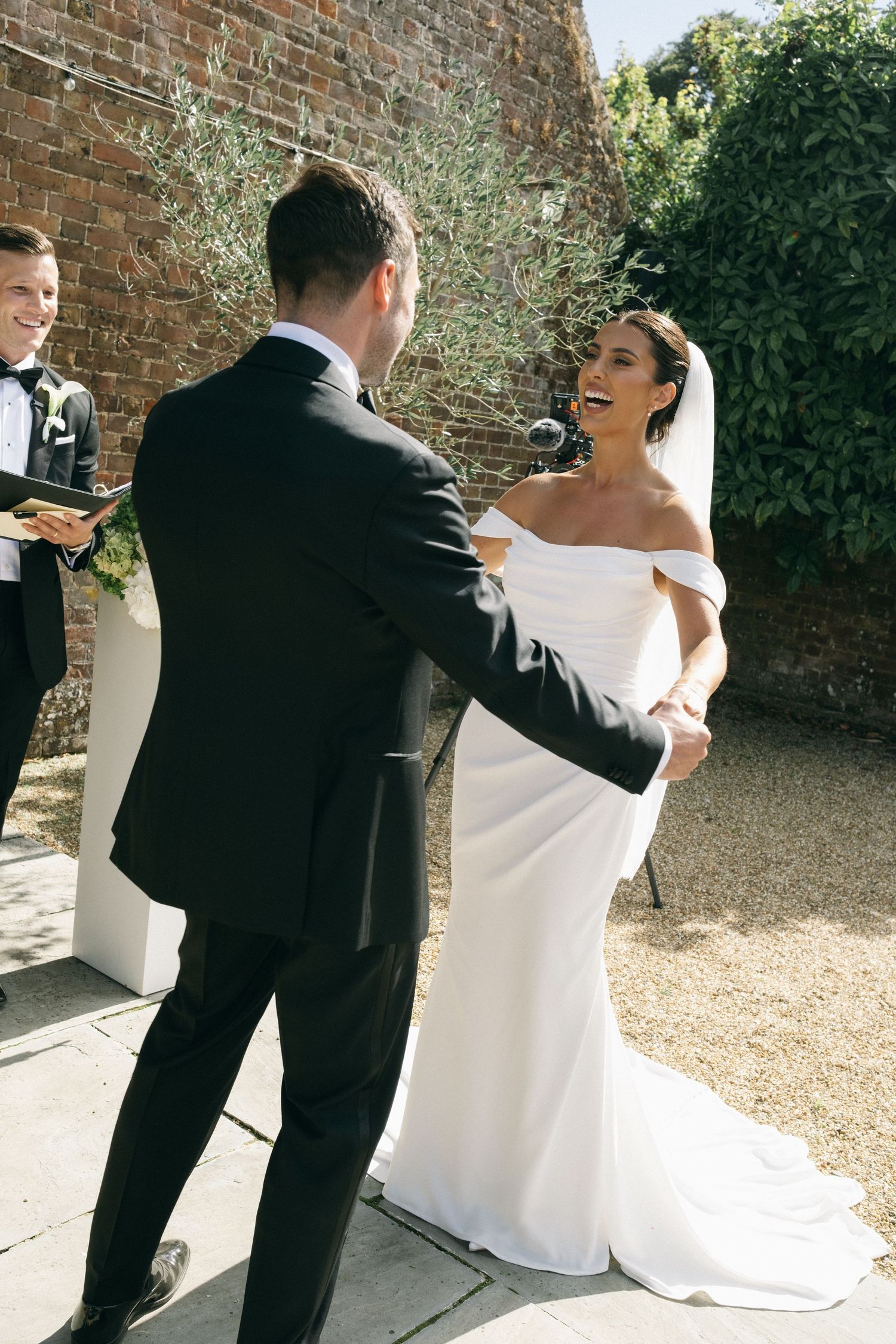 Bride and groom holding hands during outdoor wedding ceremony, bride wearing elegant off-shoulder white gown with veil.