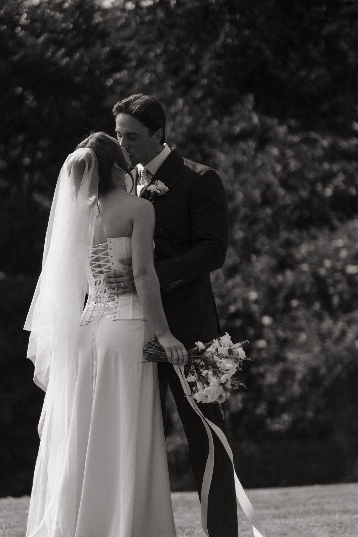 Black and white photo of a bride and groom sharing a kiss outdoors, bride holding a floral bouquet with ribbon.