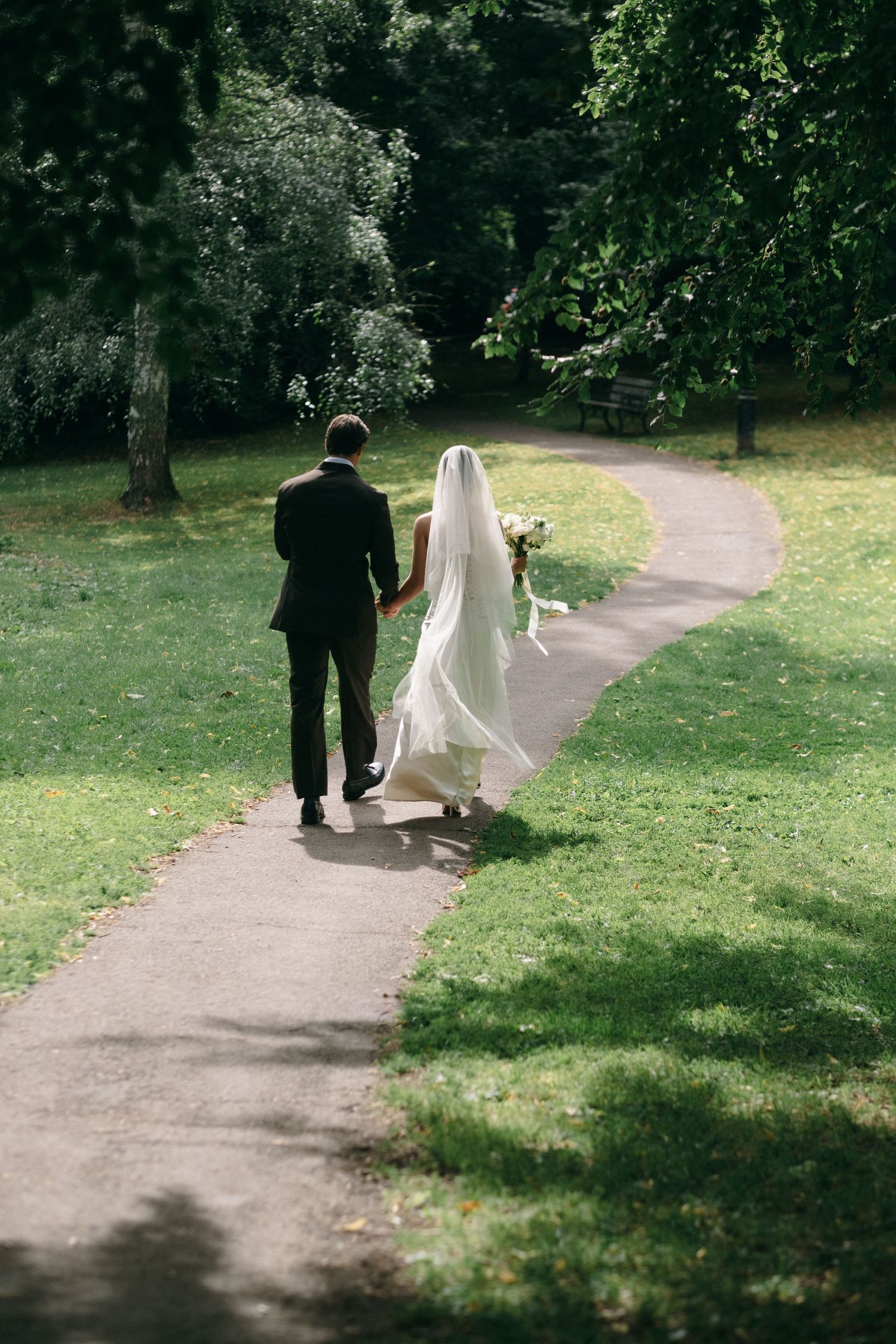 Bride and groom walking hand in hand down a sunlit park pathway, wedding veil flowing in the breeze.