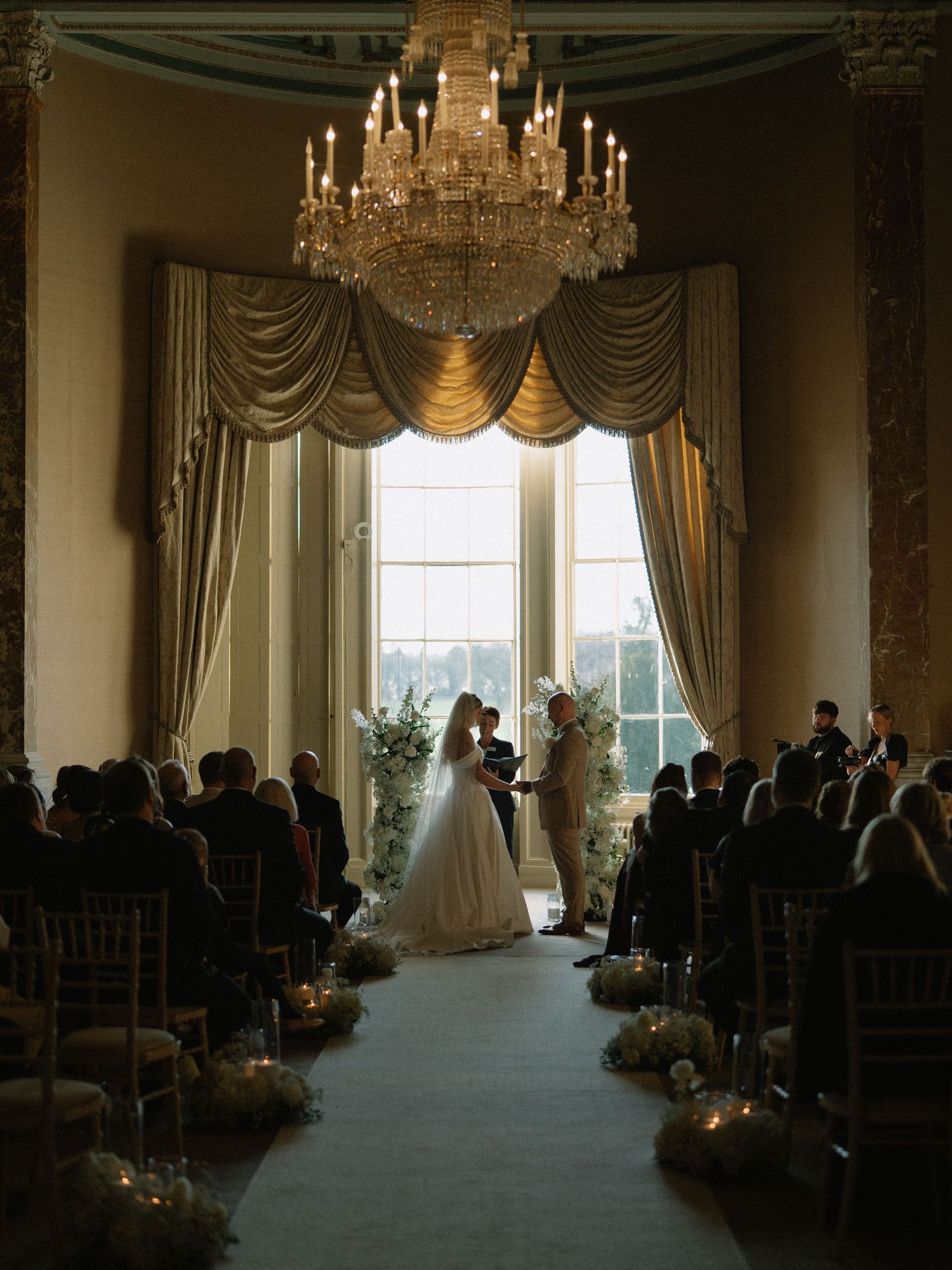 Elegant wedding ceremony in a grand ballroom with crystal chandelier, candle-lit aisle, and couple exchanging vows by tall windows.