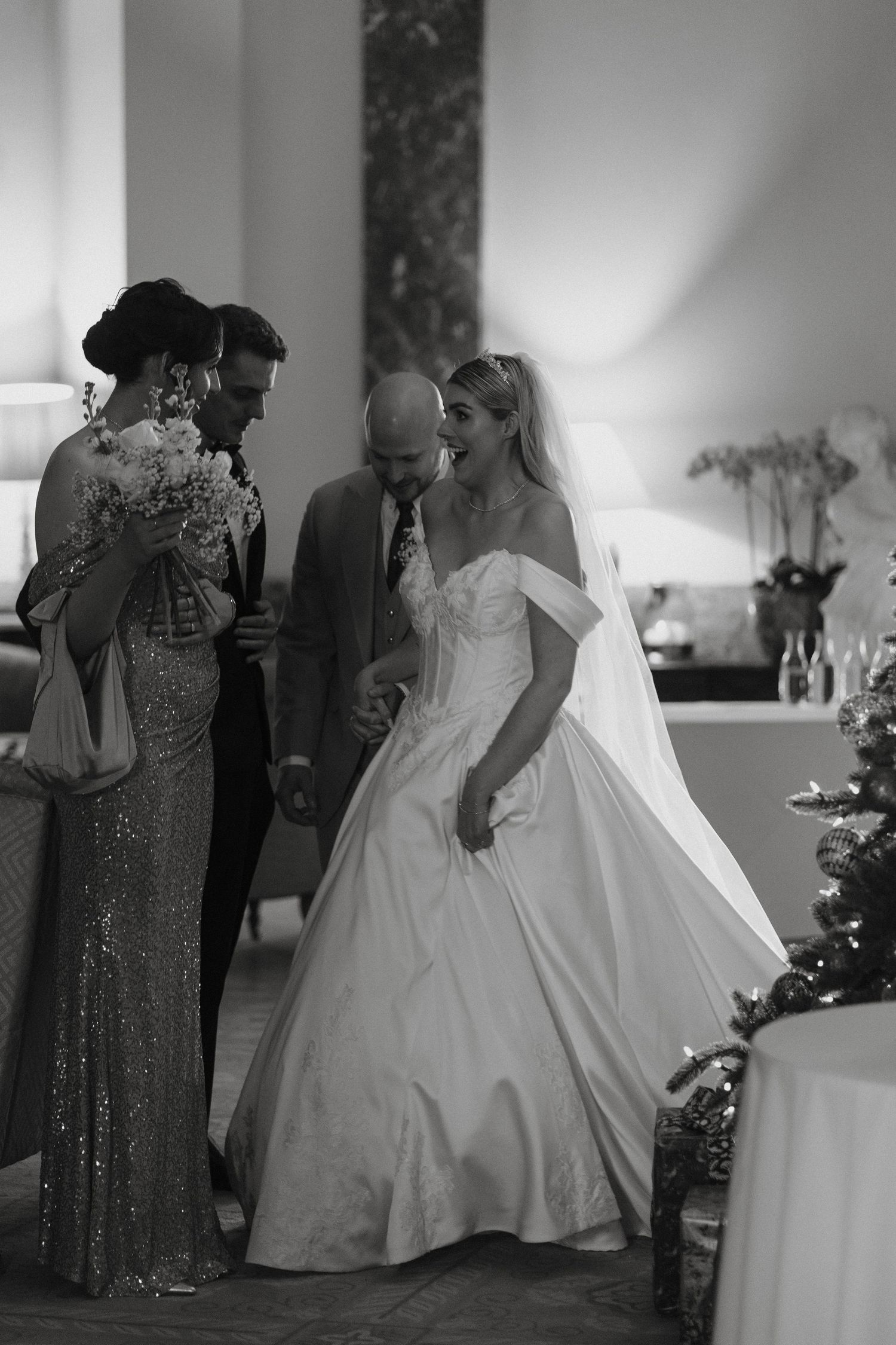 Black and white wedding photo of bride in ball gown laughing with groom and bridesmaid at indoor reception.