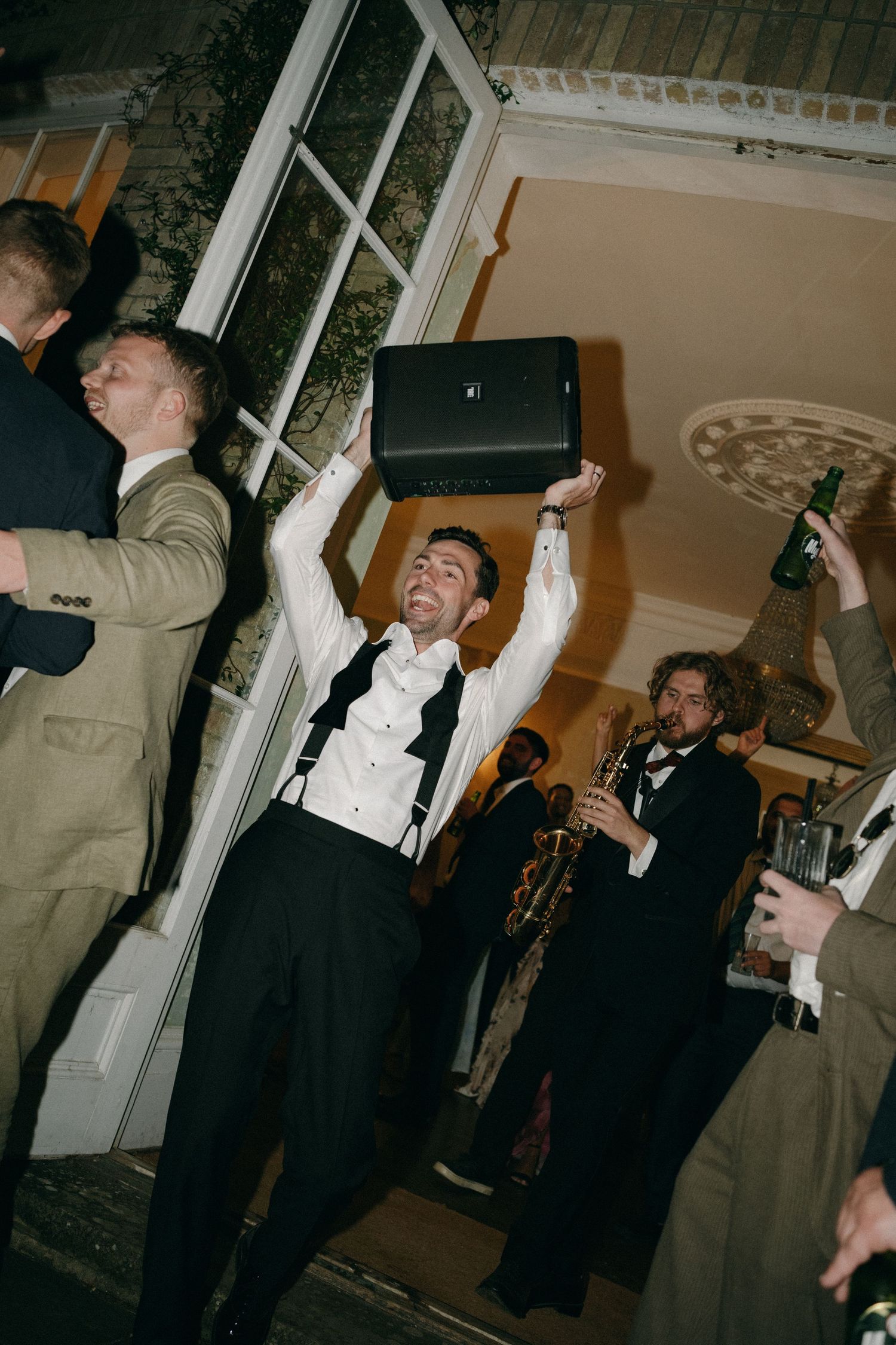 Man in tuxedo raises speaker overhead at lively wedding reception party with band playing in background.