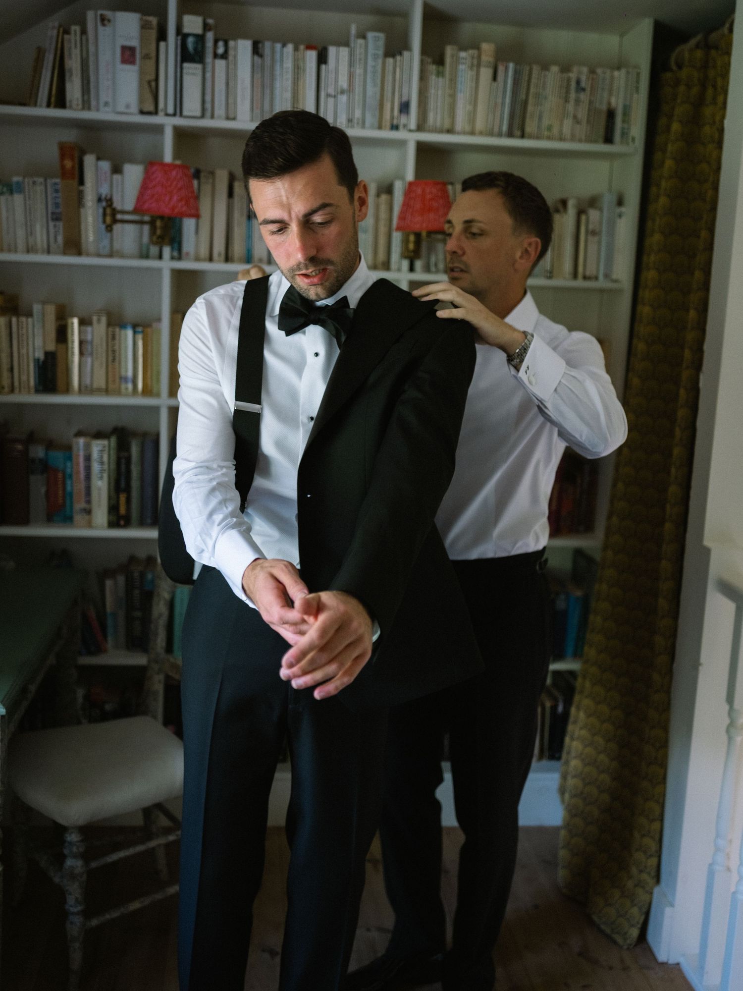 A groom in a tuxedo vest and bow tie gets help putting on his jacket in a room lined with bookshelves.