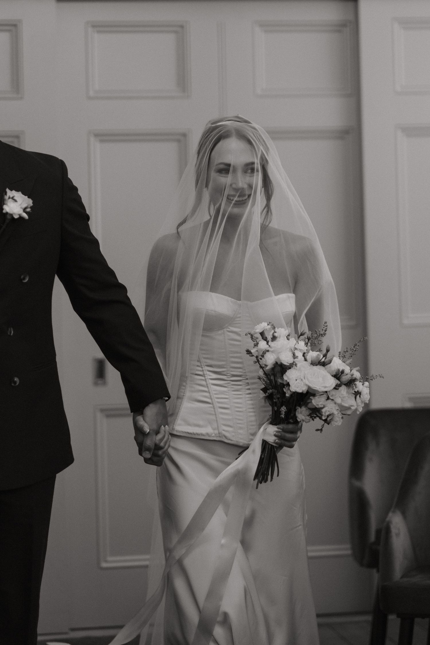 Black and white photo of a smiling bride in strapless gown and veil holding bouquet while walking with groom.