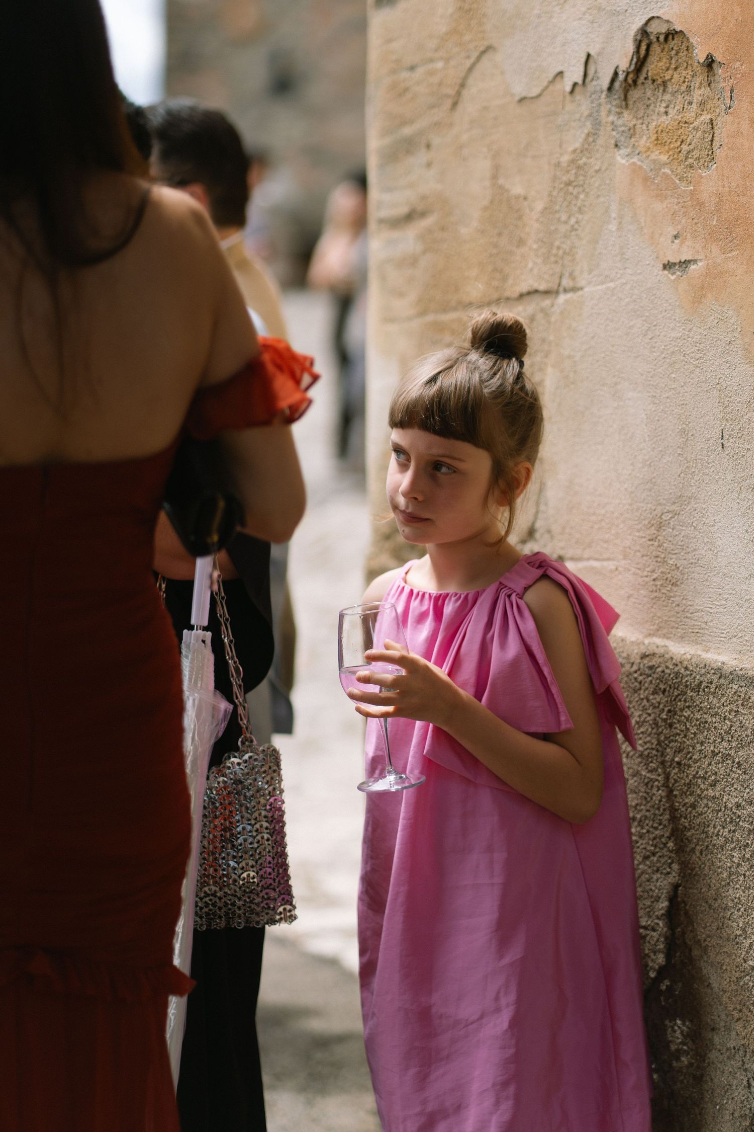 Young girl in pink dress holding champagne flute at outdoor wedding reception by stone wall.