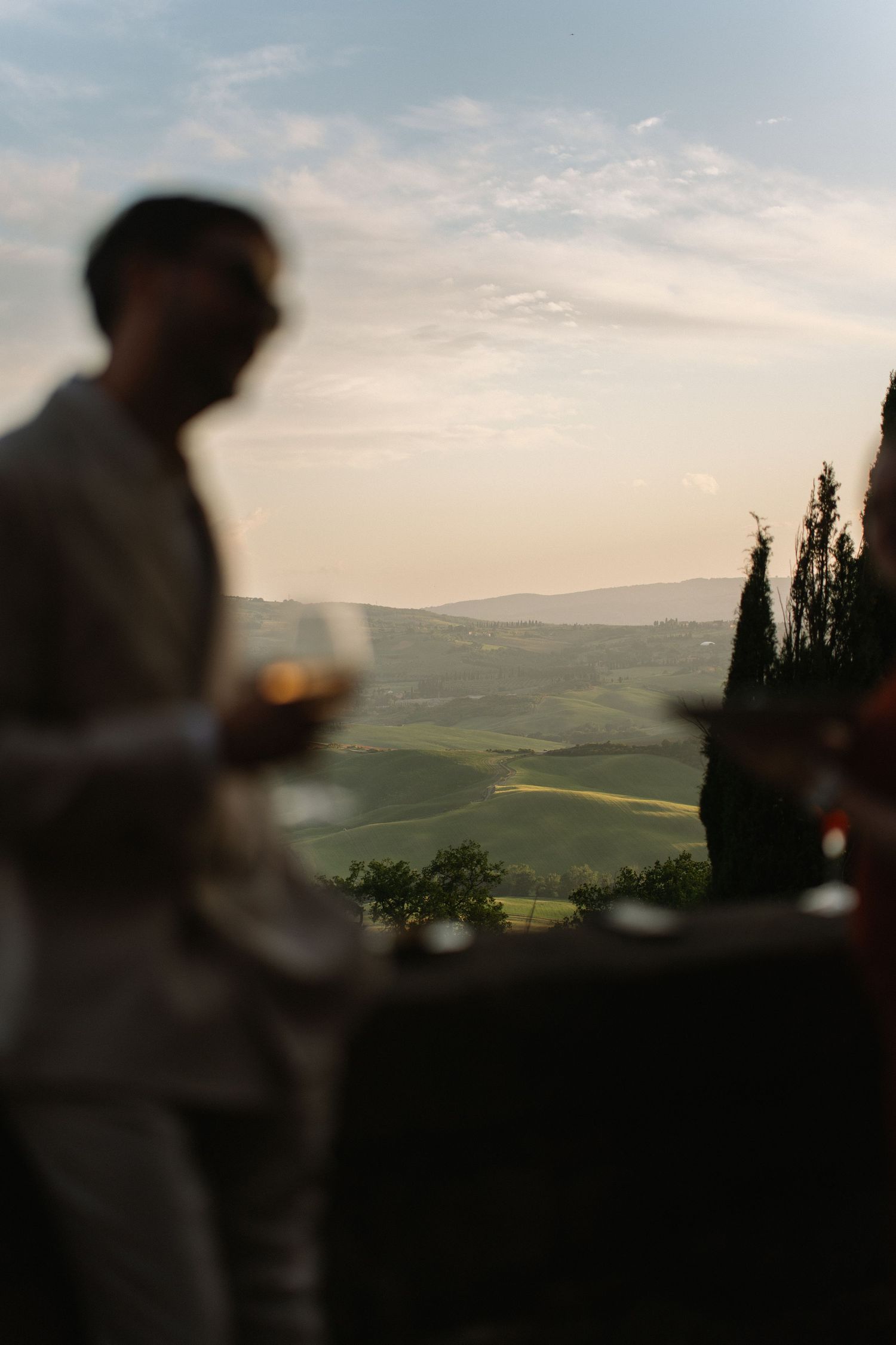 Blurred man holding wine glass at sunset overlooking rolling Tuscan hills and cypress trees.