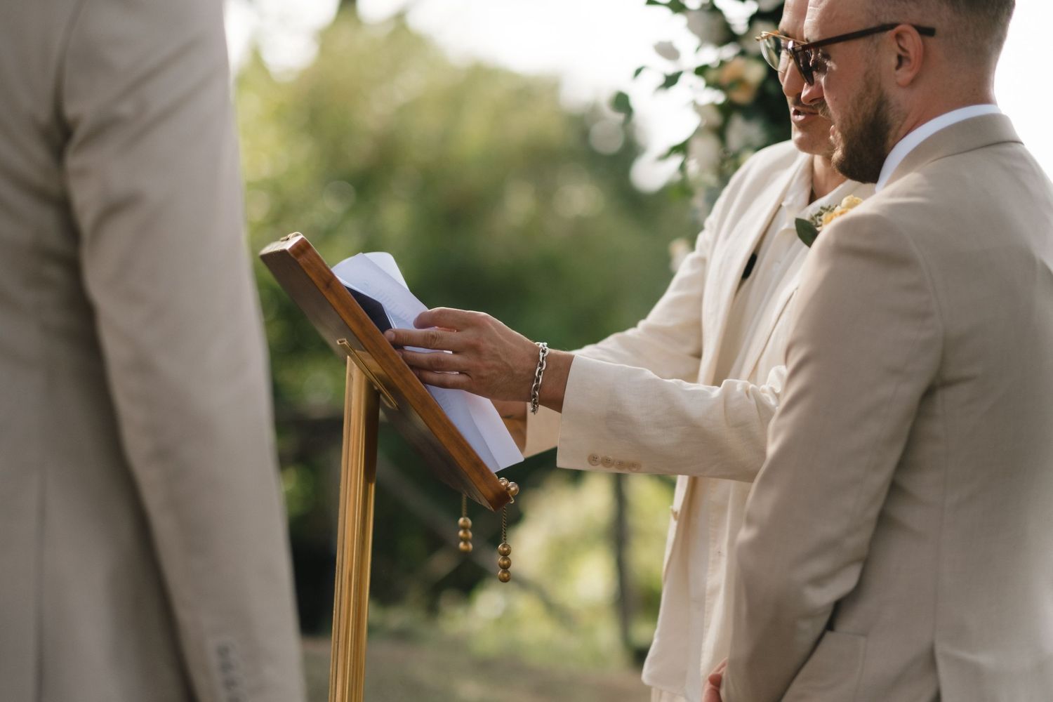 Officiant in beige suit reads from book on stand at outdoor wedding ceremony.