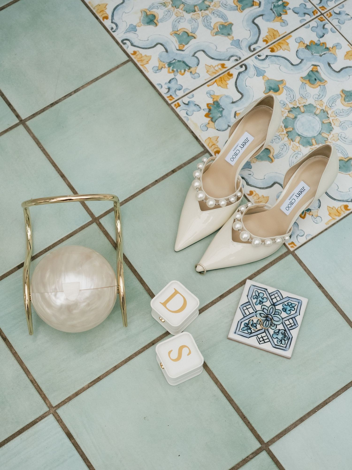 White pearl Jimmy Choo heels on teal tiles with gold clutch, monogram blocks, and decorative tile swatch.
