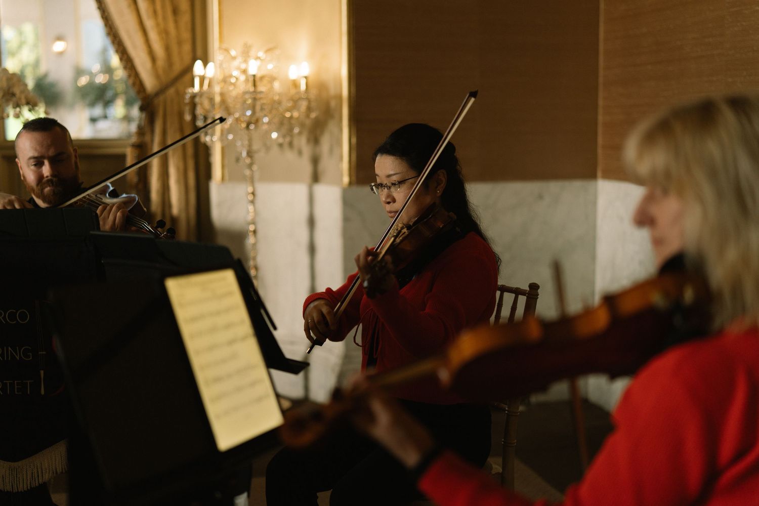 Three musicians playing violins in an elegant warmly lit room, with a woman in red at center reading sheet music.