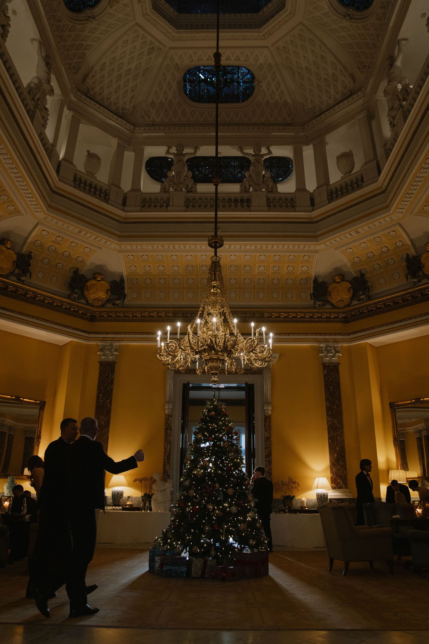 Elegant grand hall with ornate ceiling, chandelier, and decorated Christmas tree in warm golden lighting.