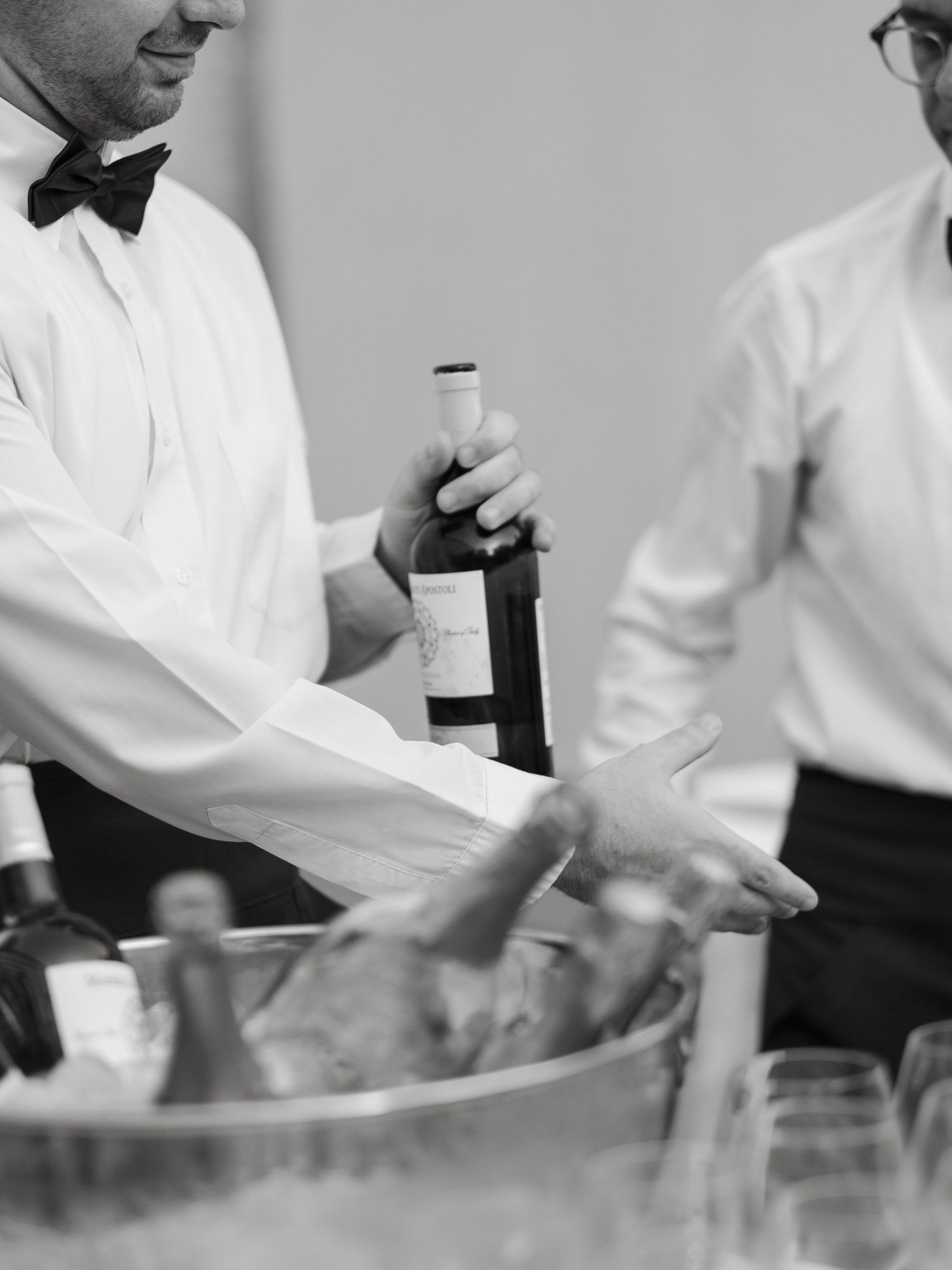 Black and white photo of a waiter in bow tie holding a wine bottle over an ice bucket at a formal event.