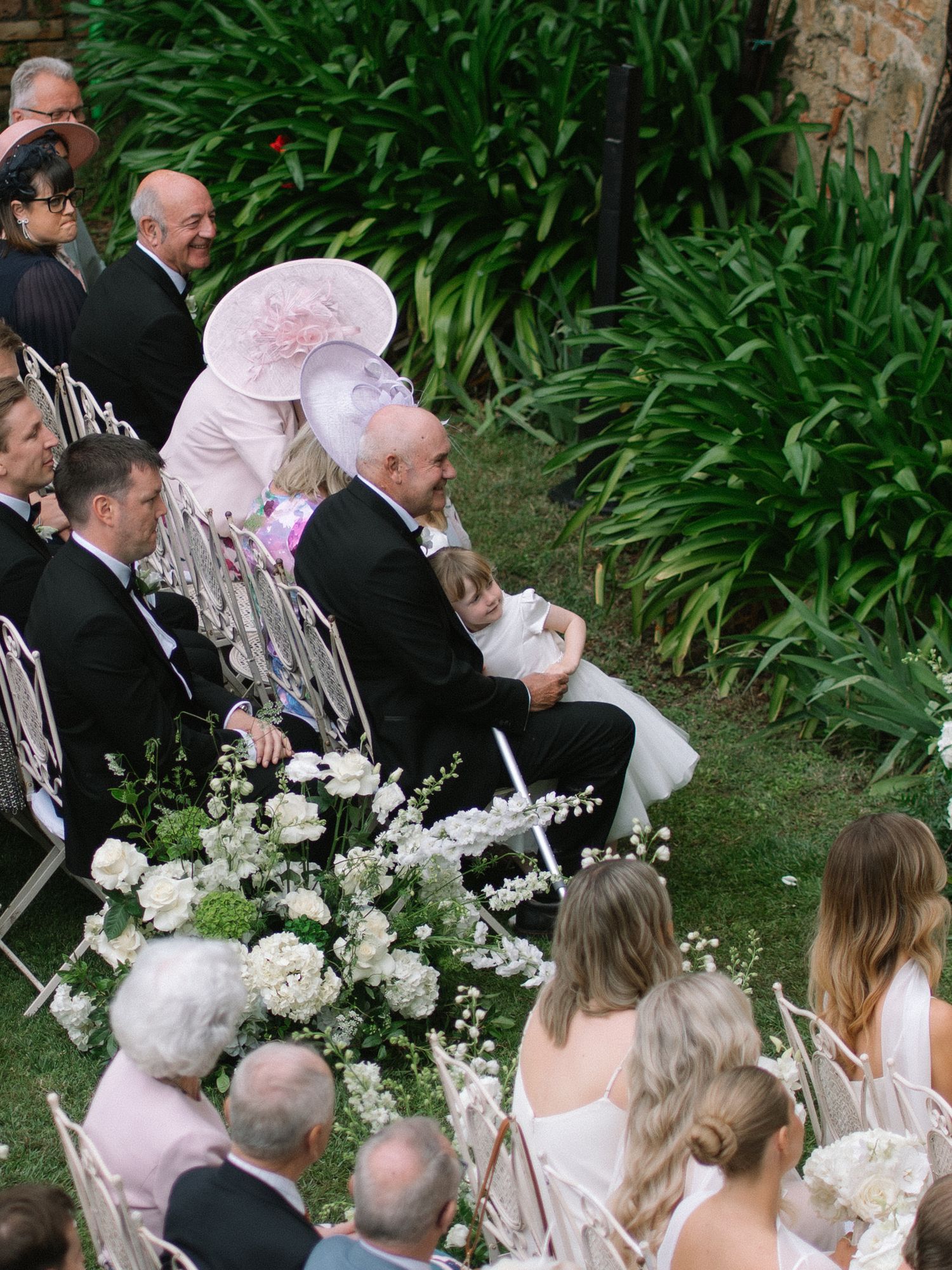 Wedding guests seated outdoors, including a woman in a large pink hat, watching the ceremony.