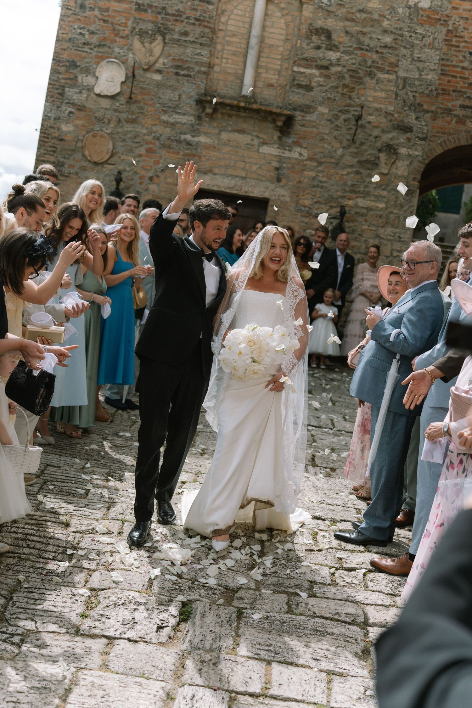 Joyful bride and groom celebrate with raised arms as guests throw confetti outside stone church.