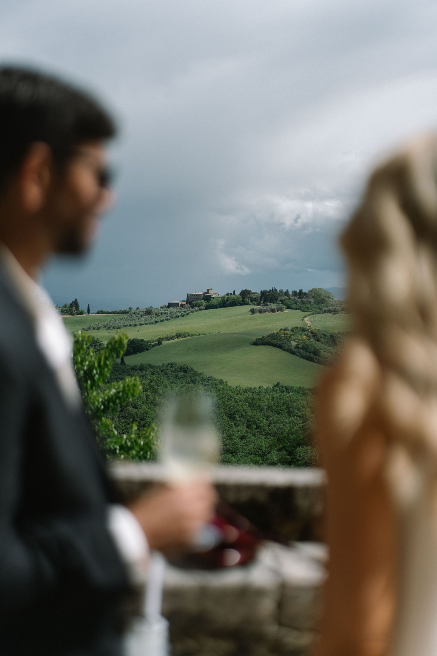 Bride and groom in soft focus holding champagne glasses with rolling Tuscan green hills in the background.