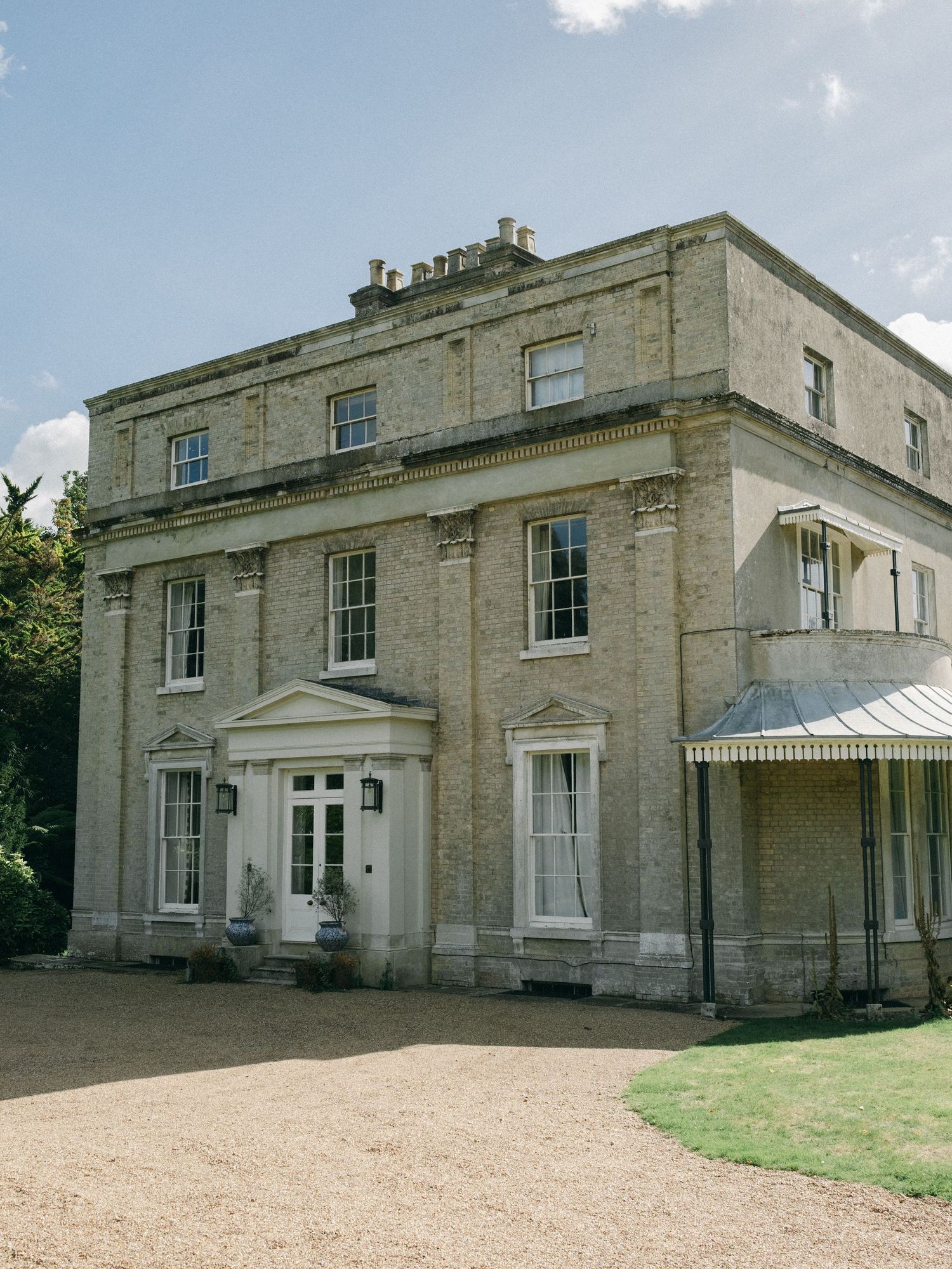 A grand Georgian country house with pale stone facade, sash windows, and gravel driveway surrounded by lush greenery.