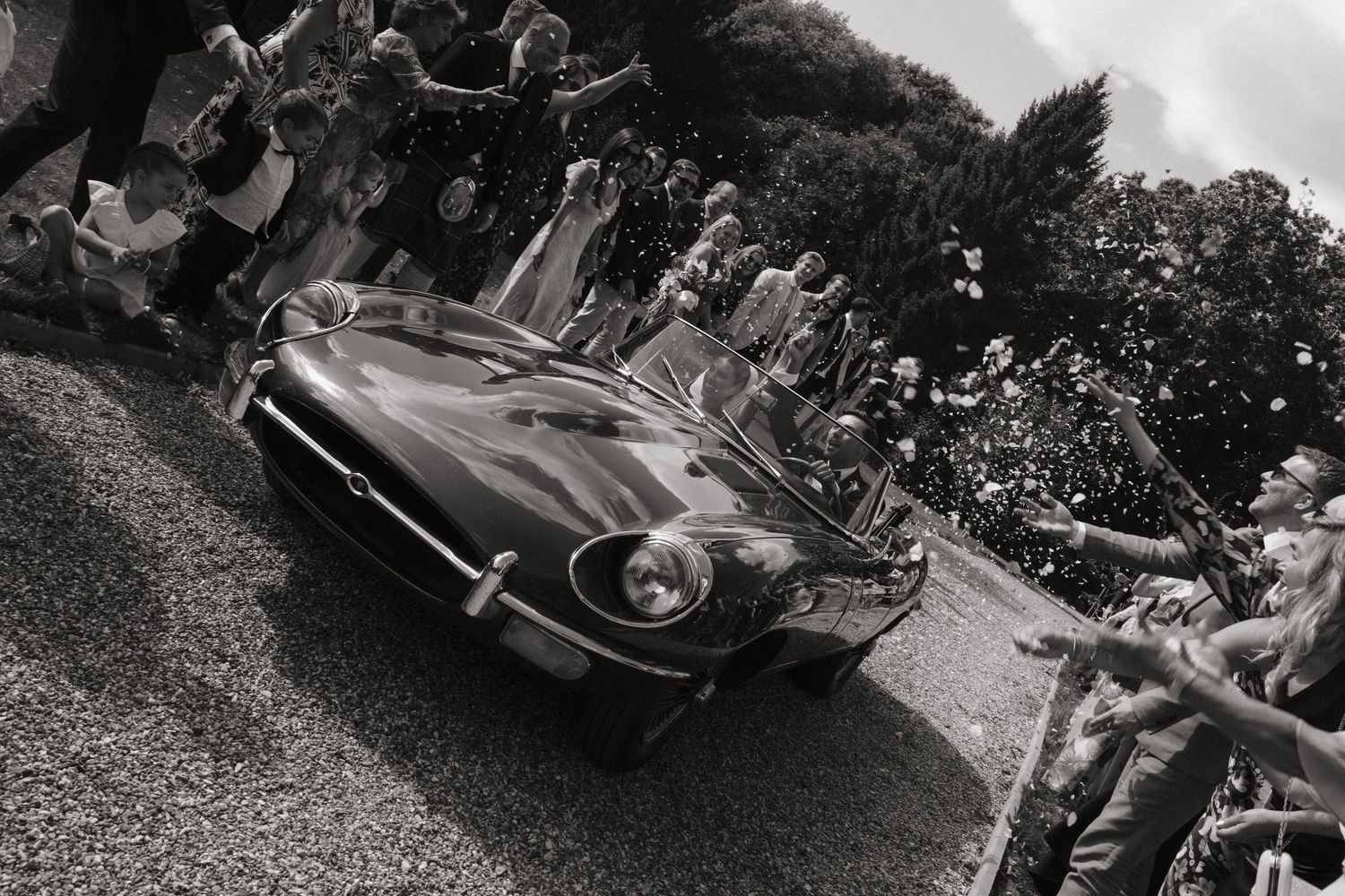Black and white photo of a classic Jaguar E-Type sports car driving past cheering crowds at a vintage car event.