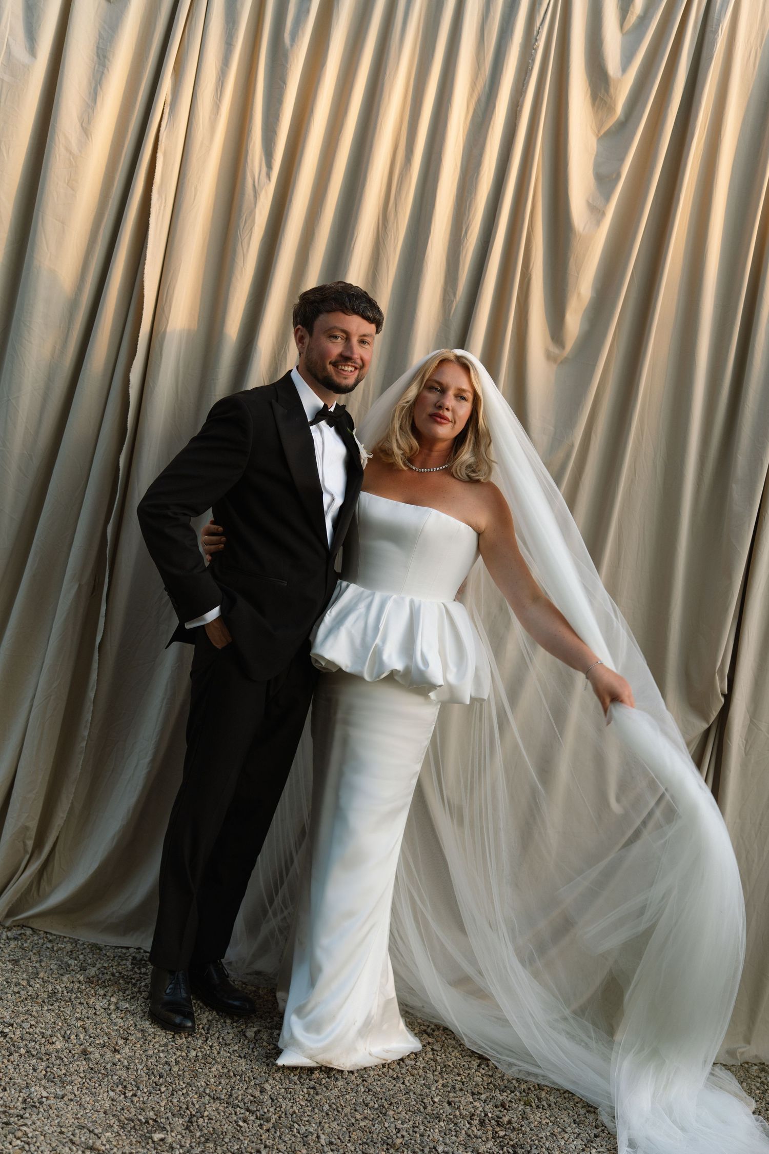 Elegant bride in strapless peplum gown with veil poses with groom in black tuxedo before draped satin backdrop.
