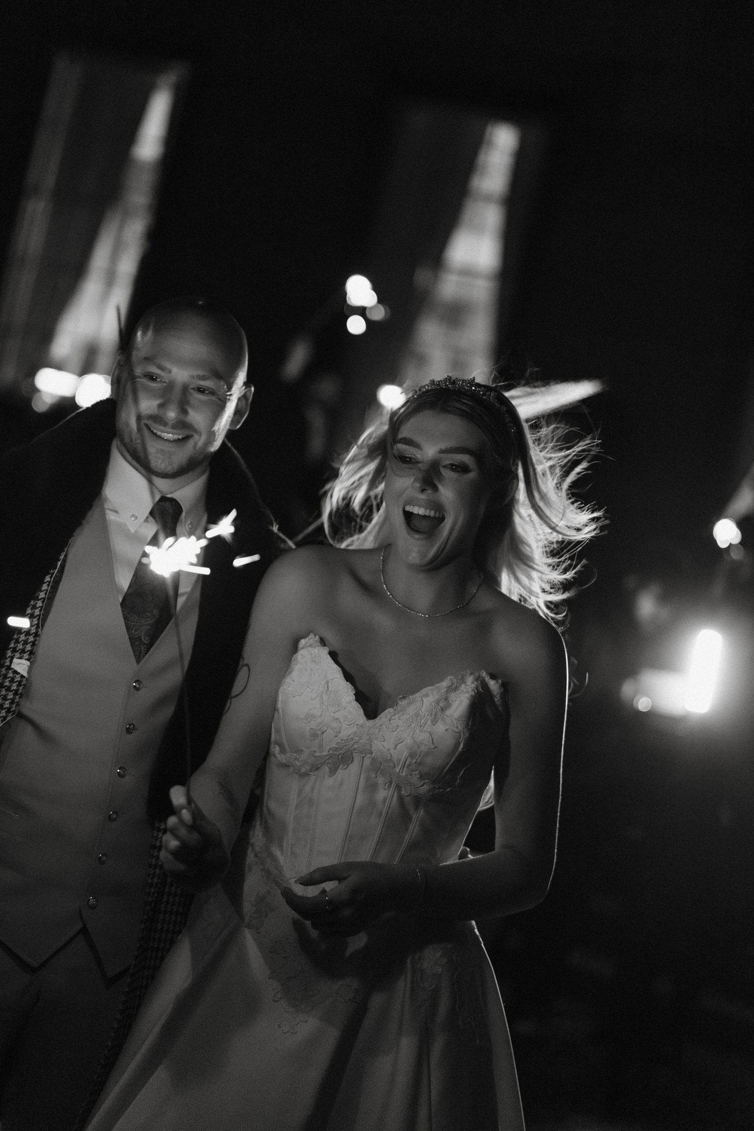 Black and white photo of a joyful bride and groom walking with sparklers at their wedding reception at night.