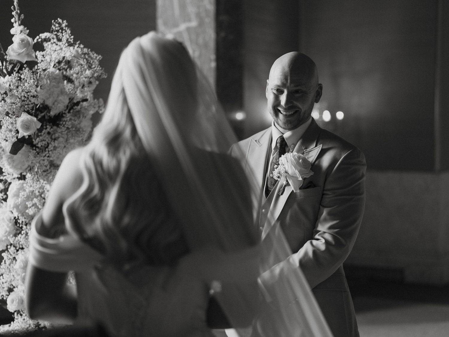Black and white wedding photo of a groom smiling at his bride in a veil near floral arrangements.