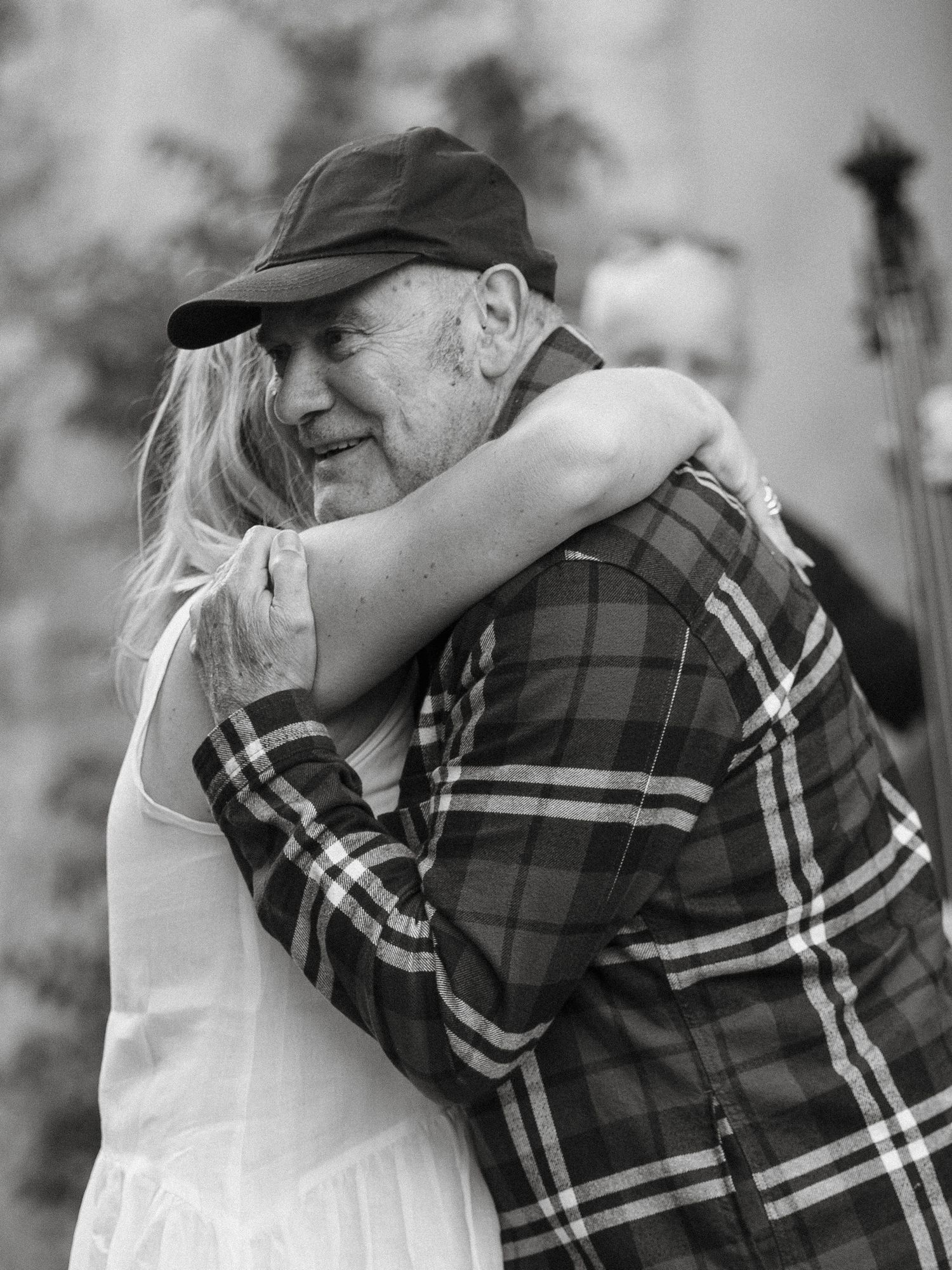 Black and white photo of an older man in plaid shirt and cap sharing a warm embrace with a young woman in white dress.
