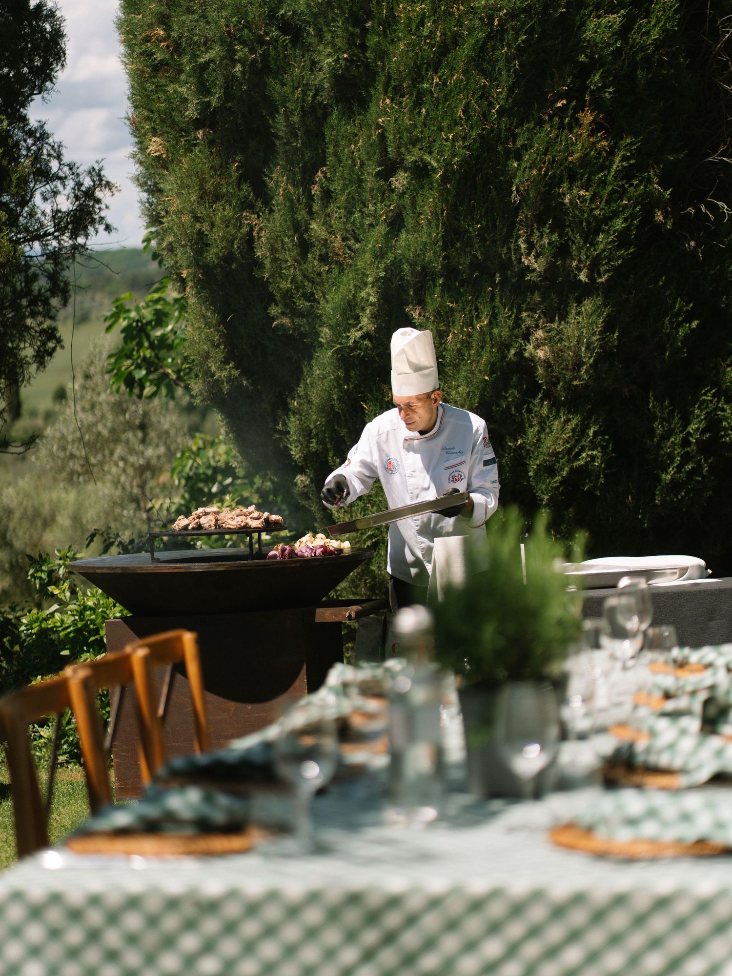 Chef in white uniform grilling meat outdoors at a Tuscan countryside wedding reception.