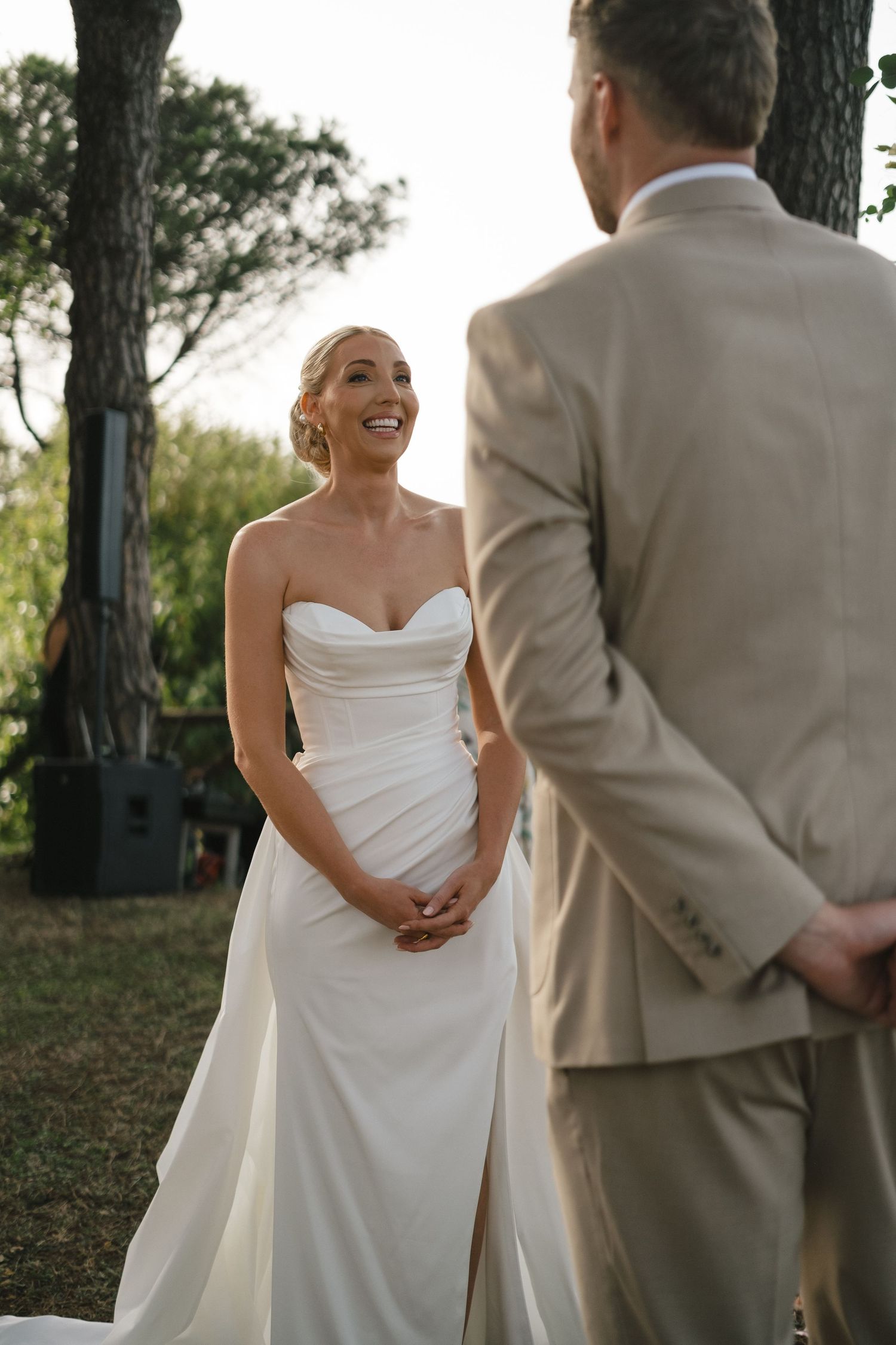 Smiling bride in strapless white gown faces groom in tan suit during outdoor wedding ceremony.