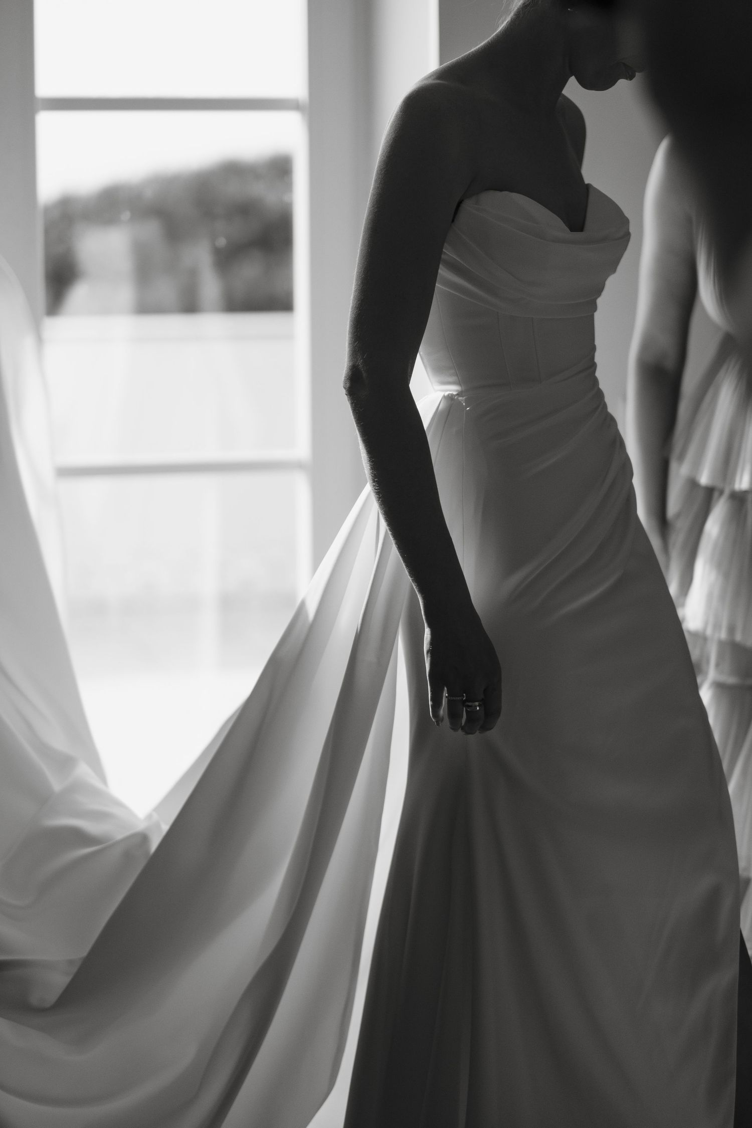 Black and white photo of a bride in a strapless wedding gown holding her dress near a bright window.
