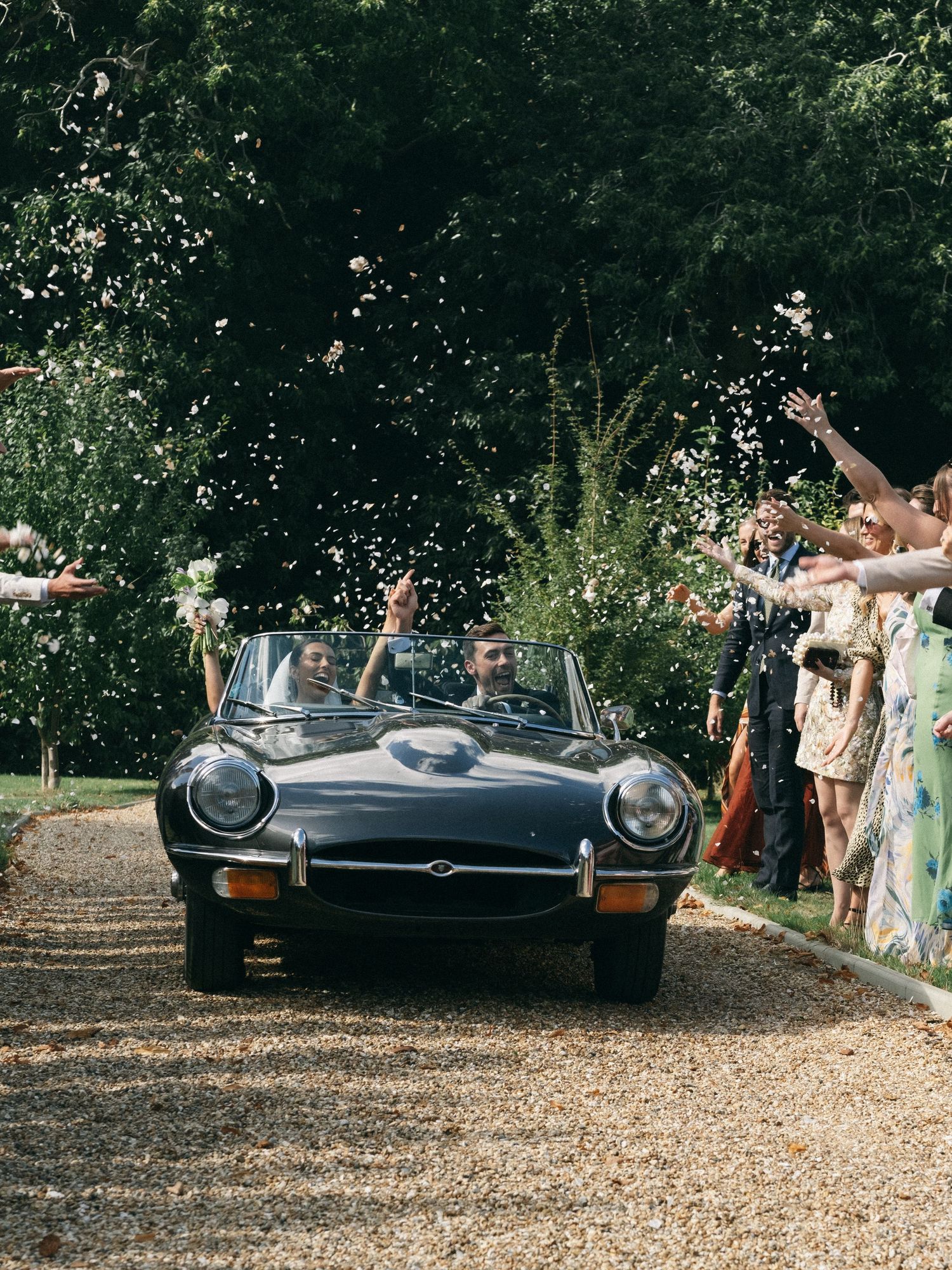Newlyweds drive a classic black Jaguar E-Type convertible through confetti-throwing wedding guests on a tree-lined gravel path.