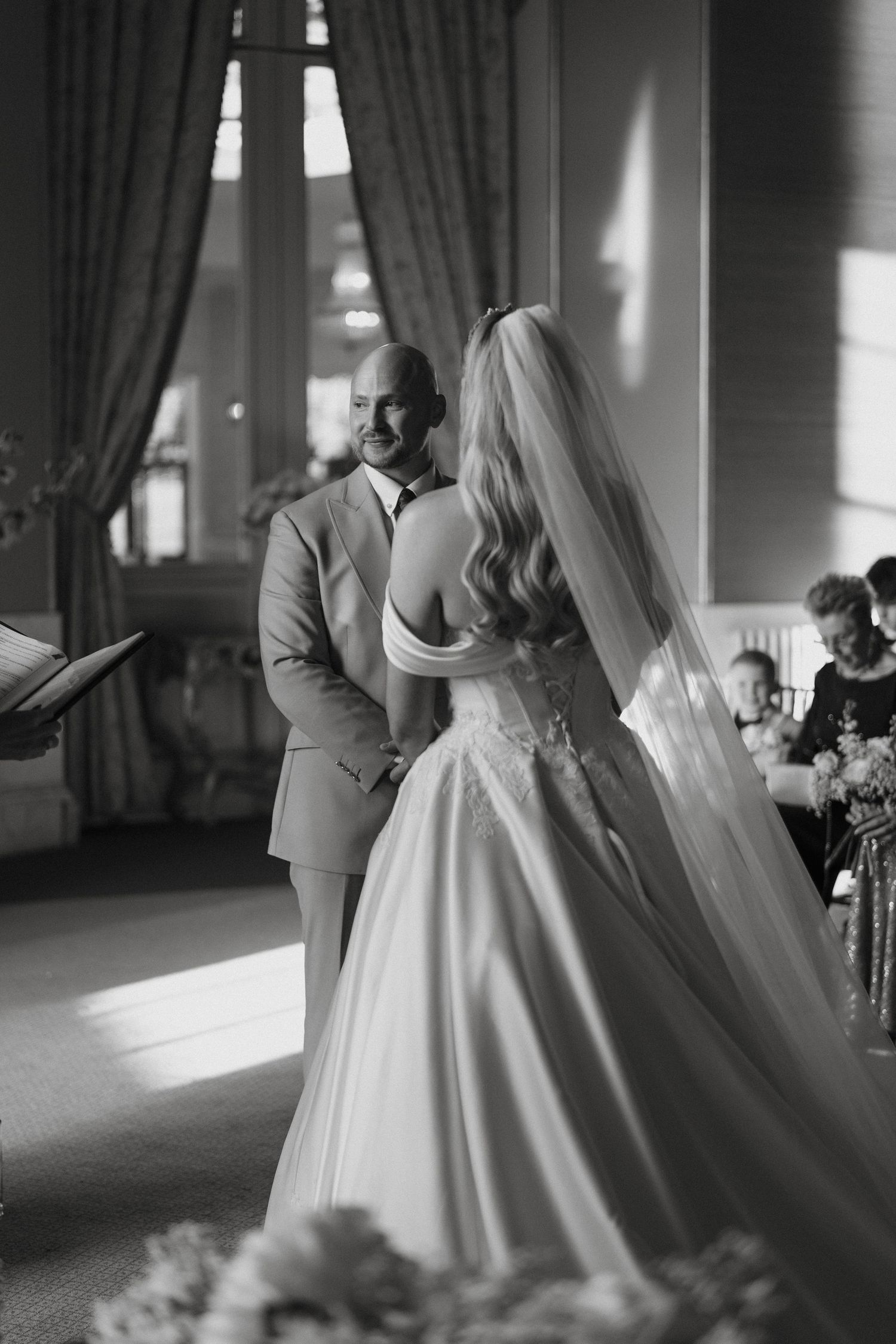 Black and white photo of a bride and groom holding hands during their wedding ceremony in an elegant venue.