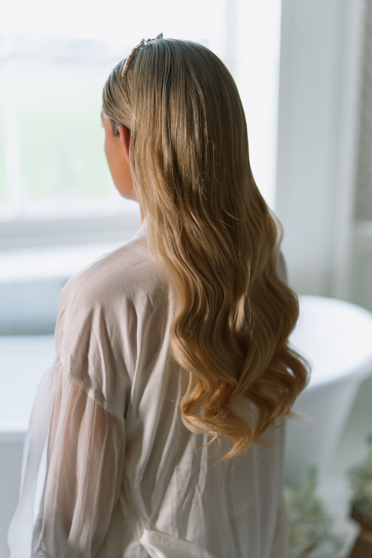 Woman with long wavy blonde hair wearing a sheer white robe, viewed from behind near a bright window.