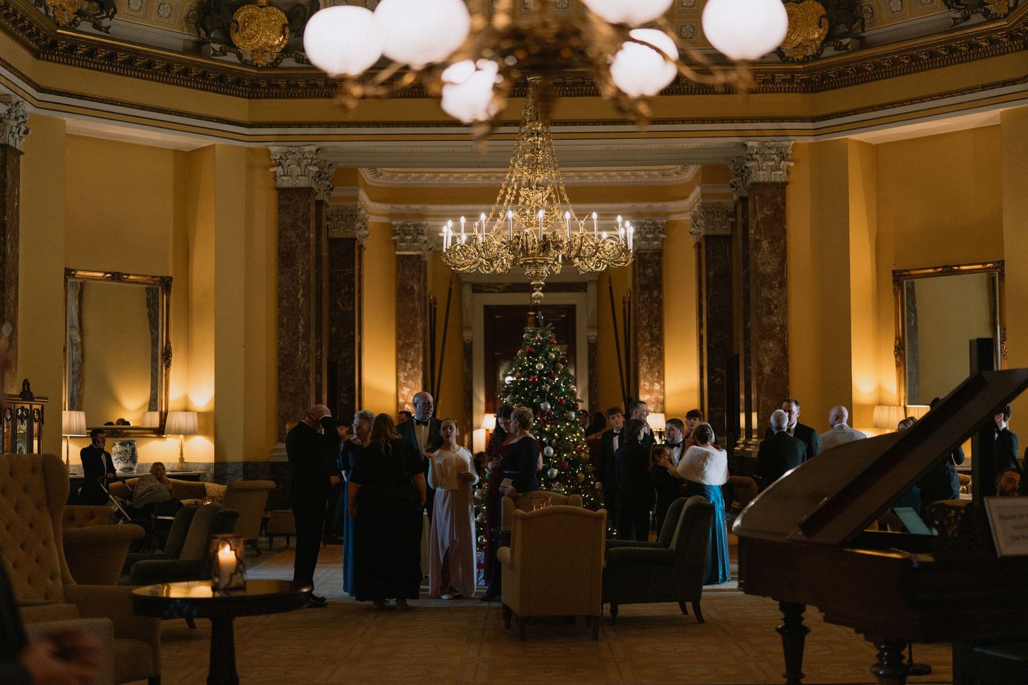 Elegant Christmas gathering in a grand ballroom with chandelier, decorated tree, and guests in formal attire.