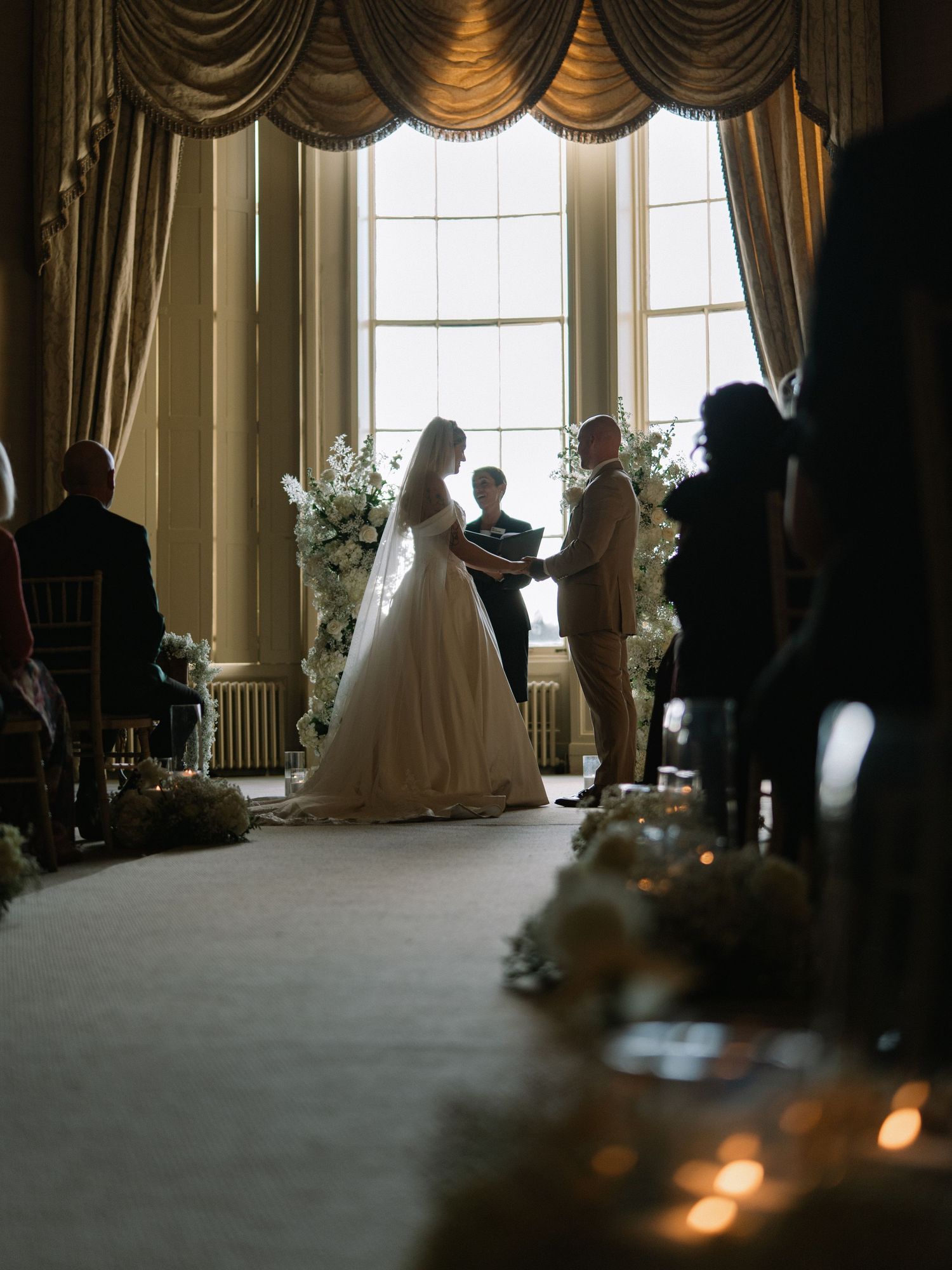 Elegant wedding ceremony in a grand ballroom with bride and groom exchanging vows before large windows with gold drapes.