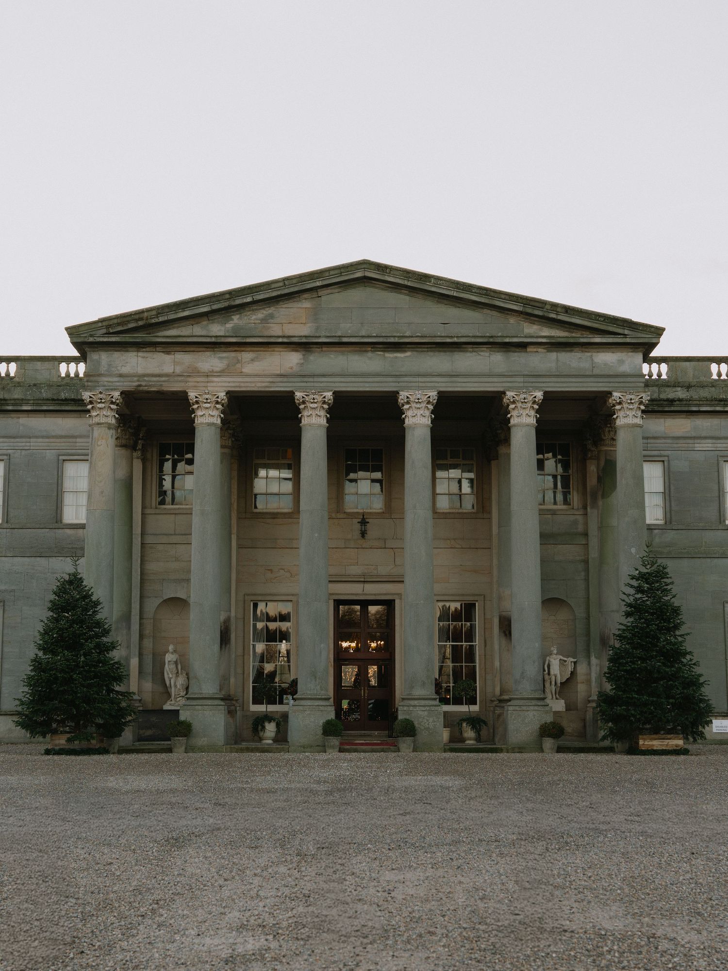 Grand neoclassical mansion with Corinthian columns, triangular pediment, and flanking evergreen trees on a gravel forecourt.