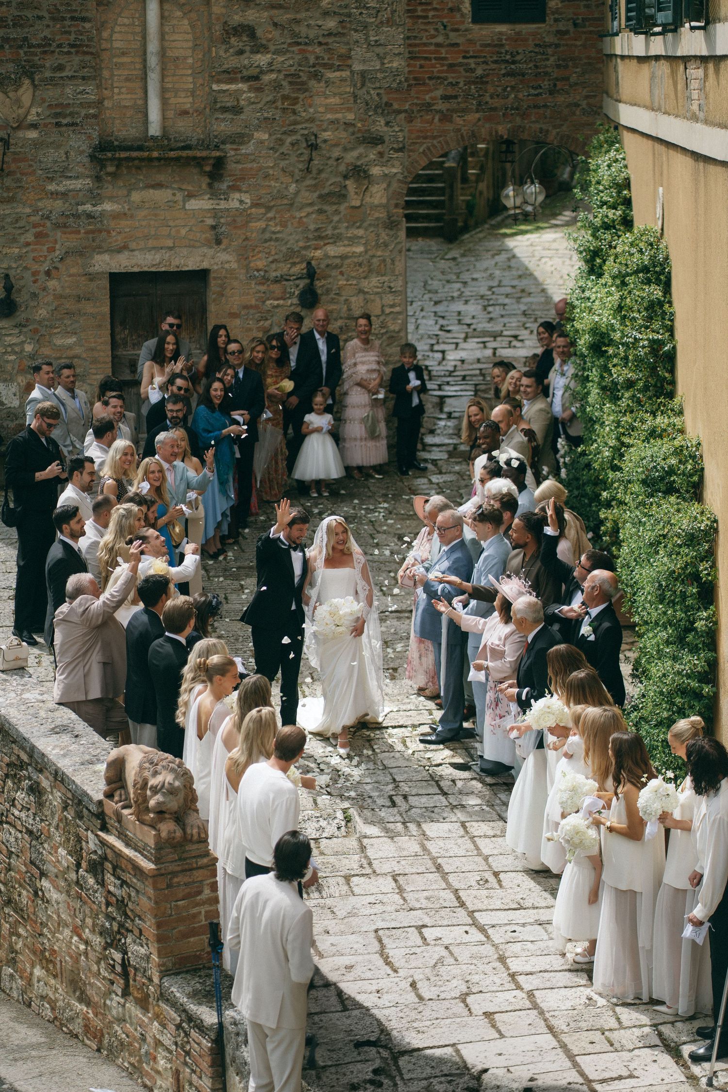 Aerial view of wedding couple walking cobblestone path lined with guests tossing confetti at historic venue.