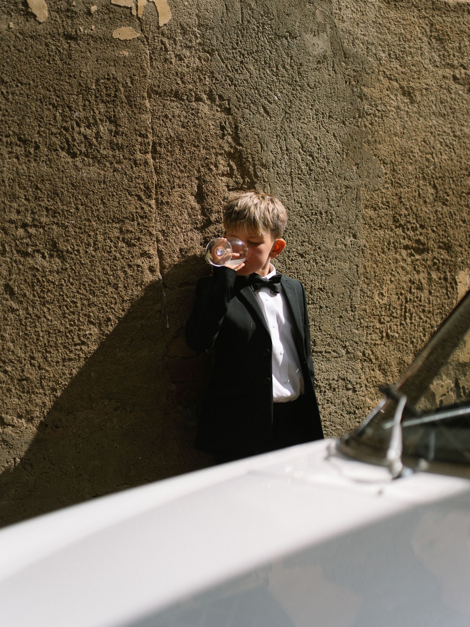 Young boy in a black tuxedo drinking from a glass while leaning against a rustic stone wall in Italy.