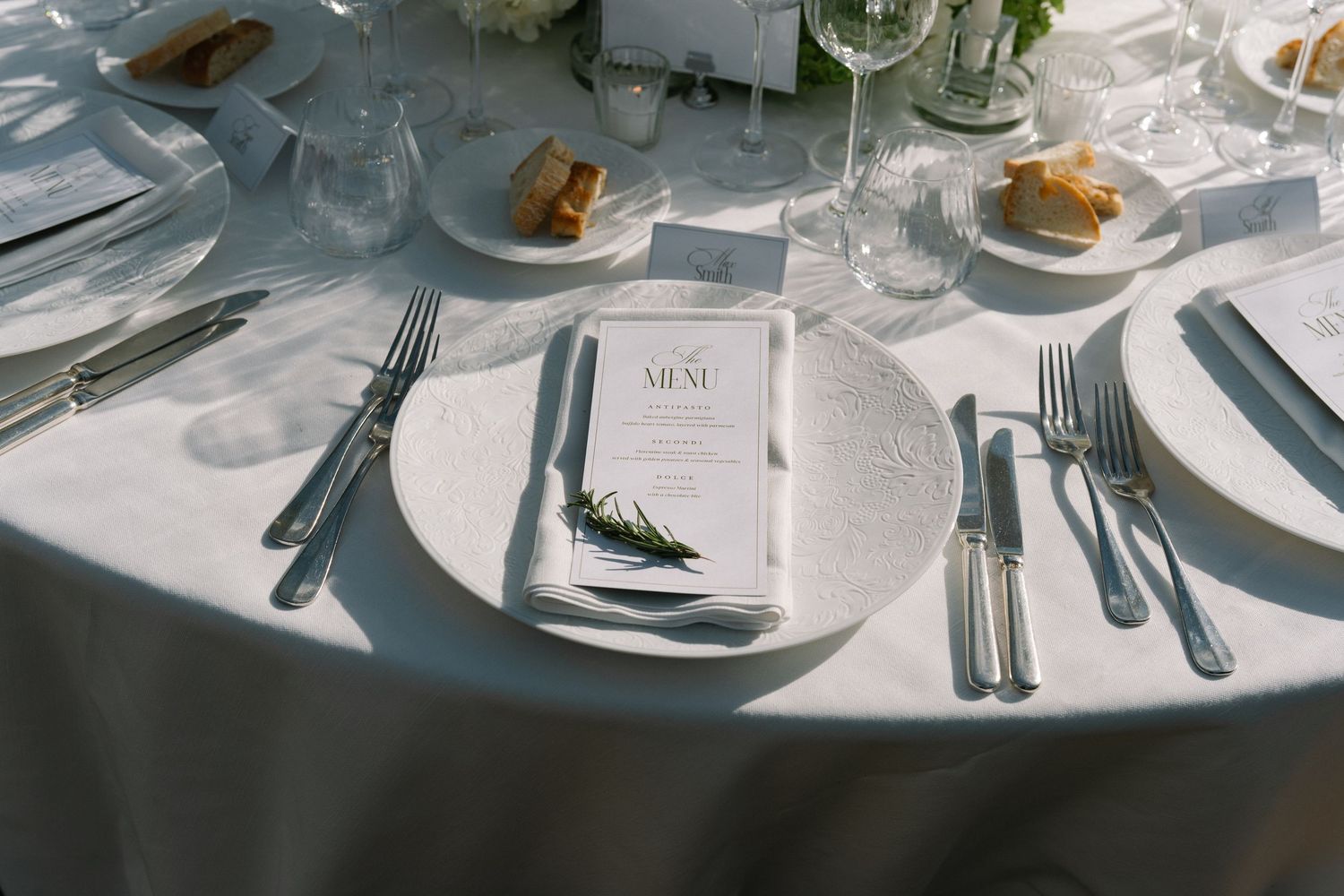 Wedding reception round table setting with white menu card, fresh rosemary sprig, silverware, and white linen tablecloth.