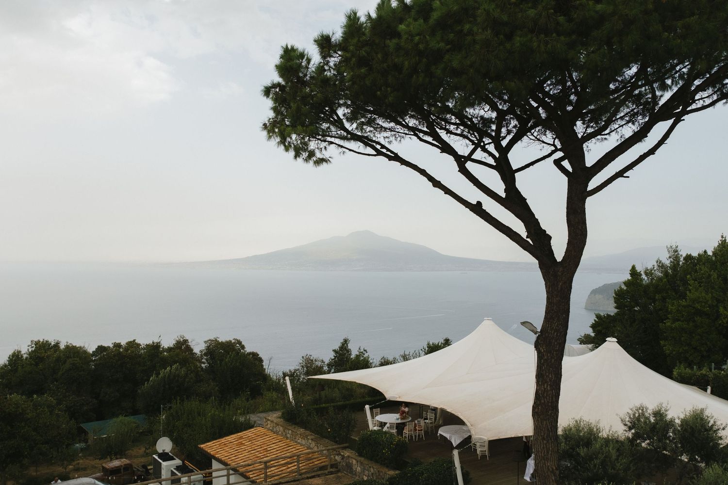 Scenic coastal view with white event tents, lush trees, and misty mountain silhouette over calm sea waters.
