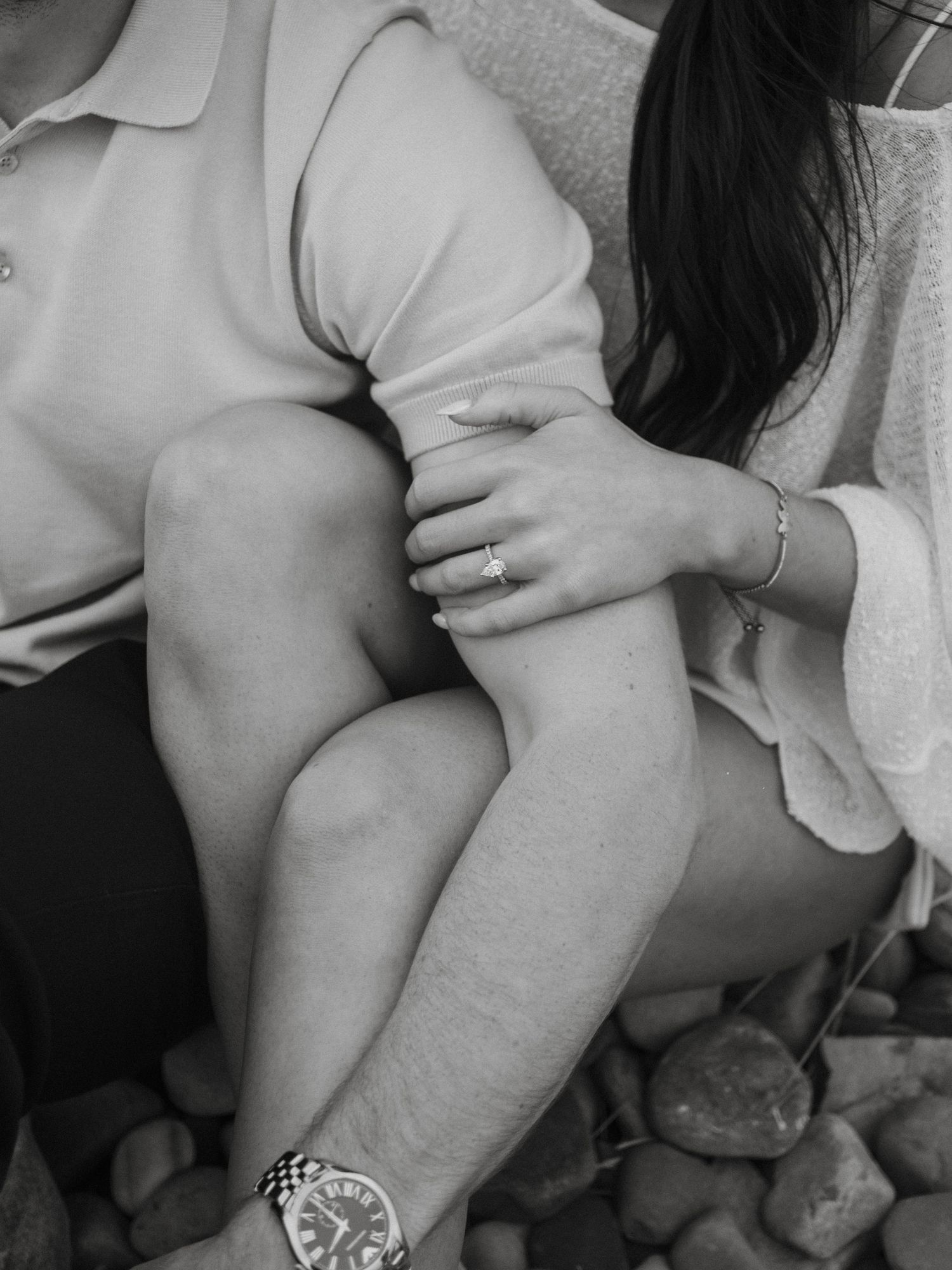 Black and white close-up of couple sitting on rocks showing engagement ring.