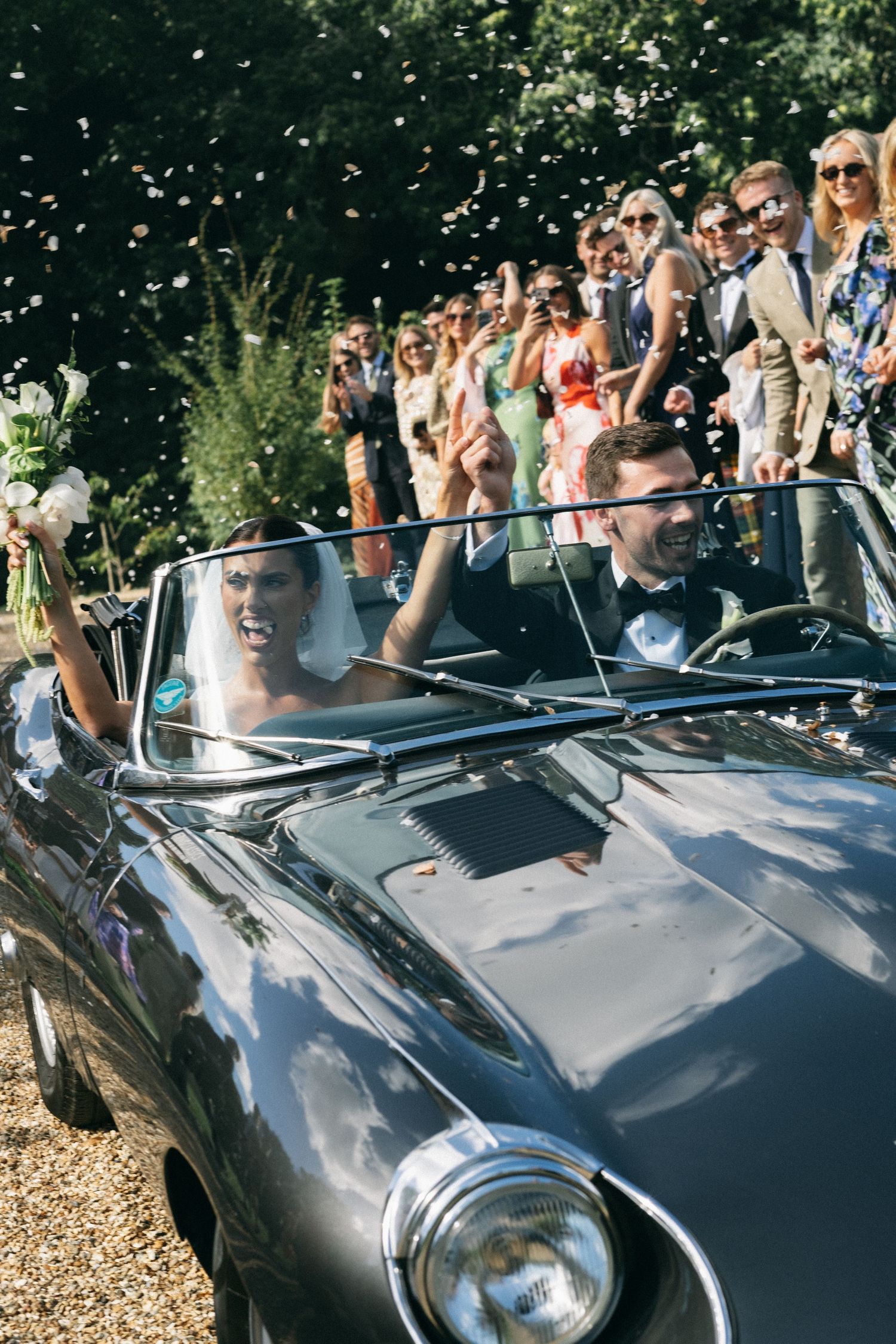 Newlyweds celebrate in a classic vintage convertible car as wedding guests throw confetti outdoors.