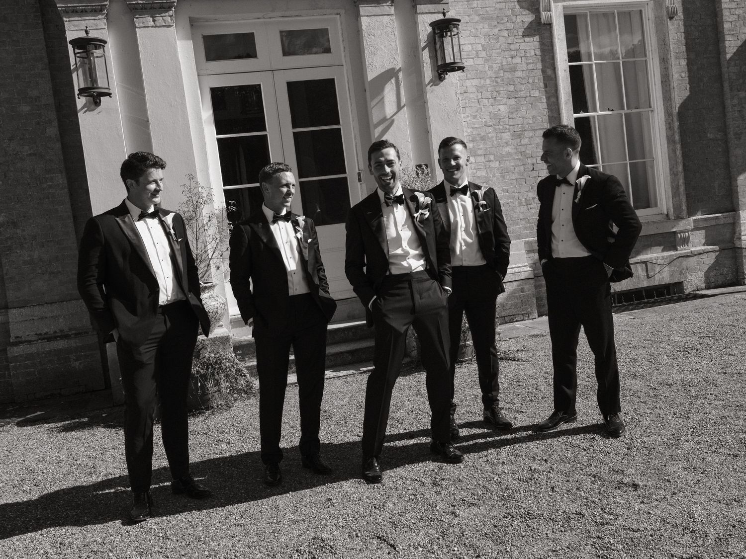 Five groomsmen in black tuxedos pose outdoors in front of an elegant manor house in a black and white photo.