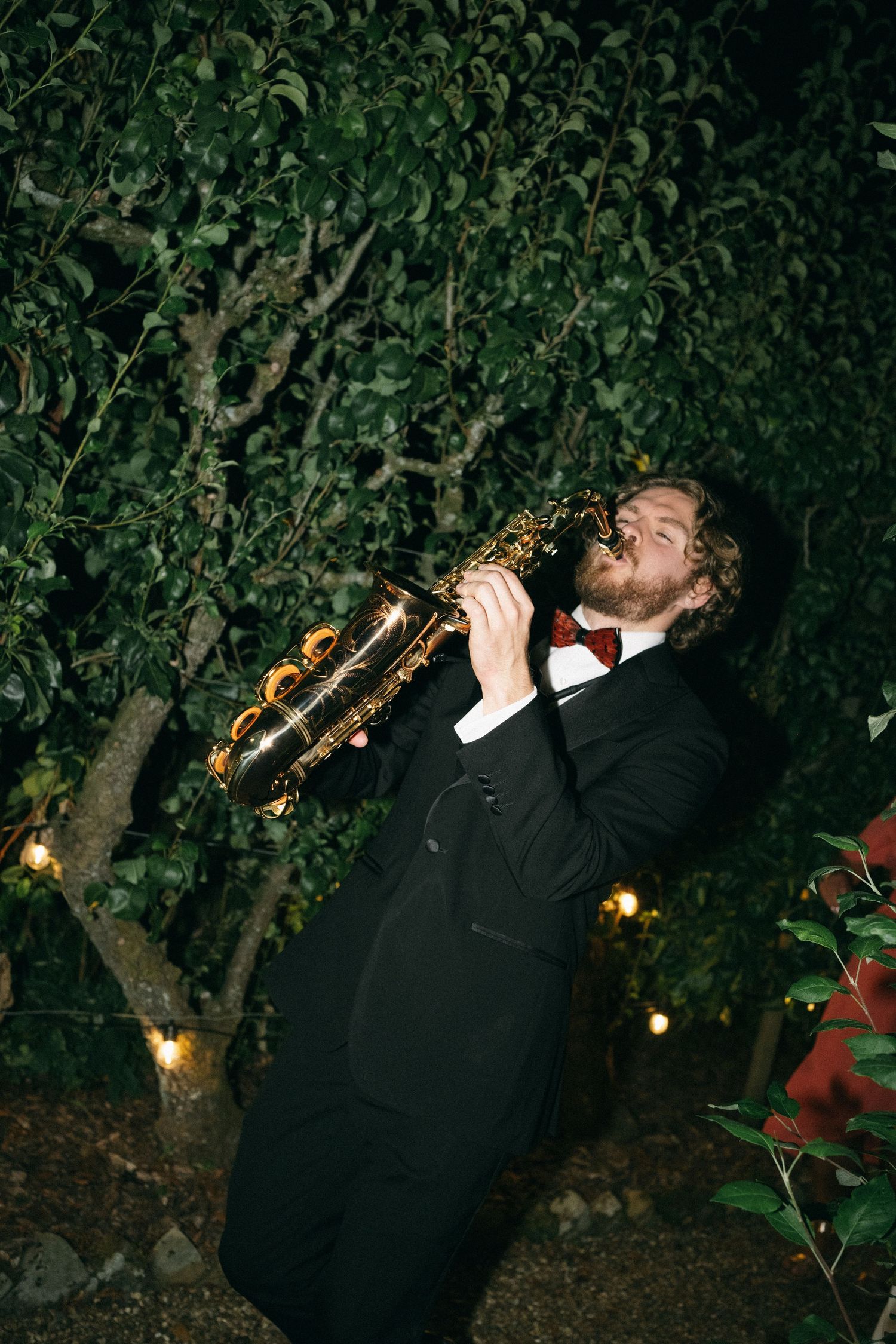 A man in a tuxedo and bow tie passionately plays a saxophone outdoors at night among lush green trees with string lights.