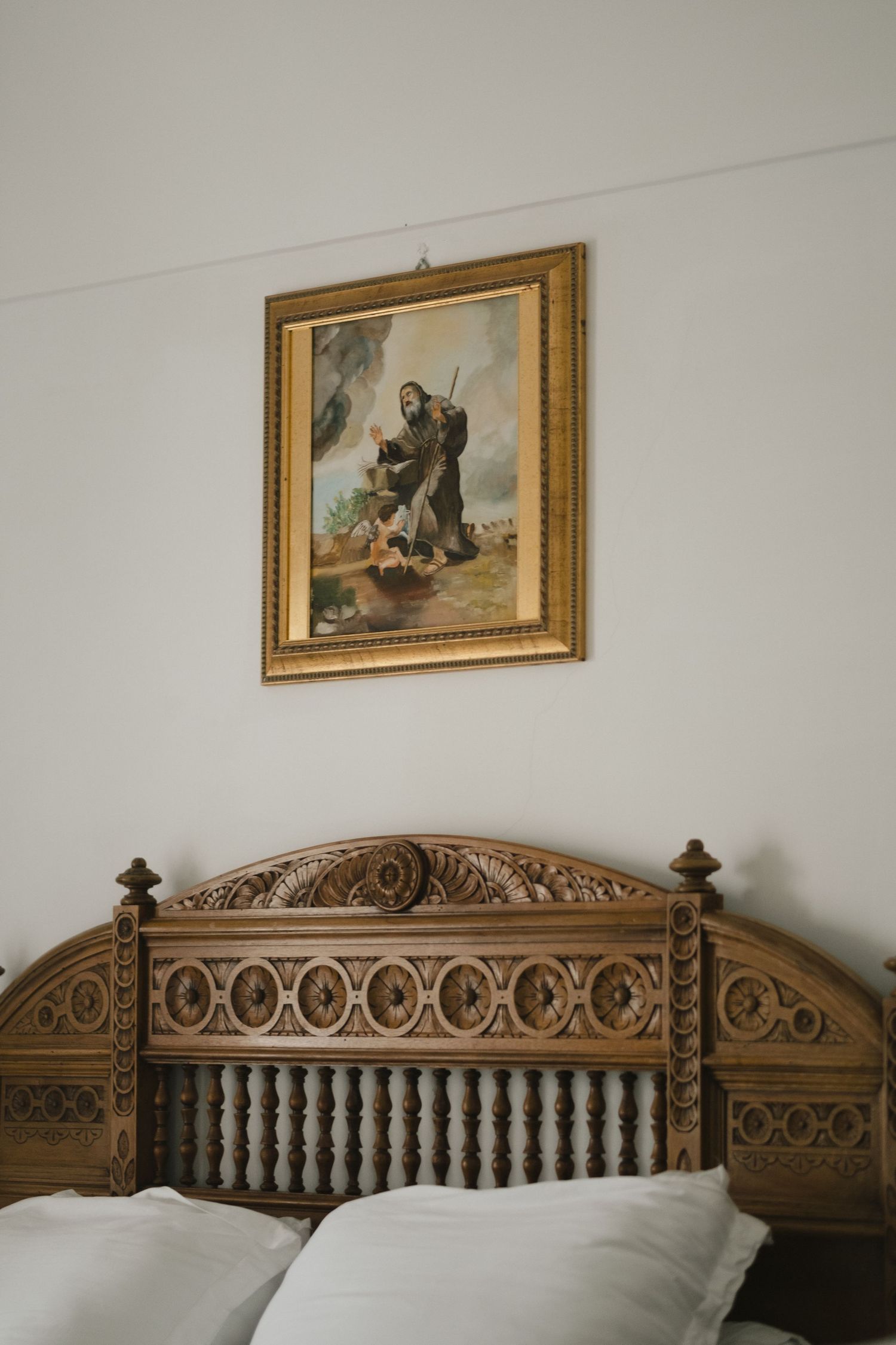 Ornate carved wooden bed headboard with white pillows beneath a gold-framed religious painting on a white wall.