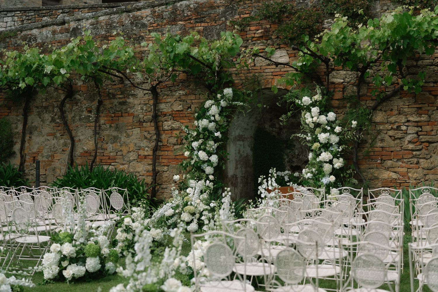 Outdoor wedding ceremony setup with white chairs, floral arch, and lush vines against an ancient brick wall.