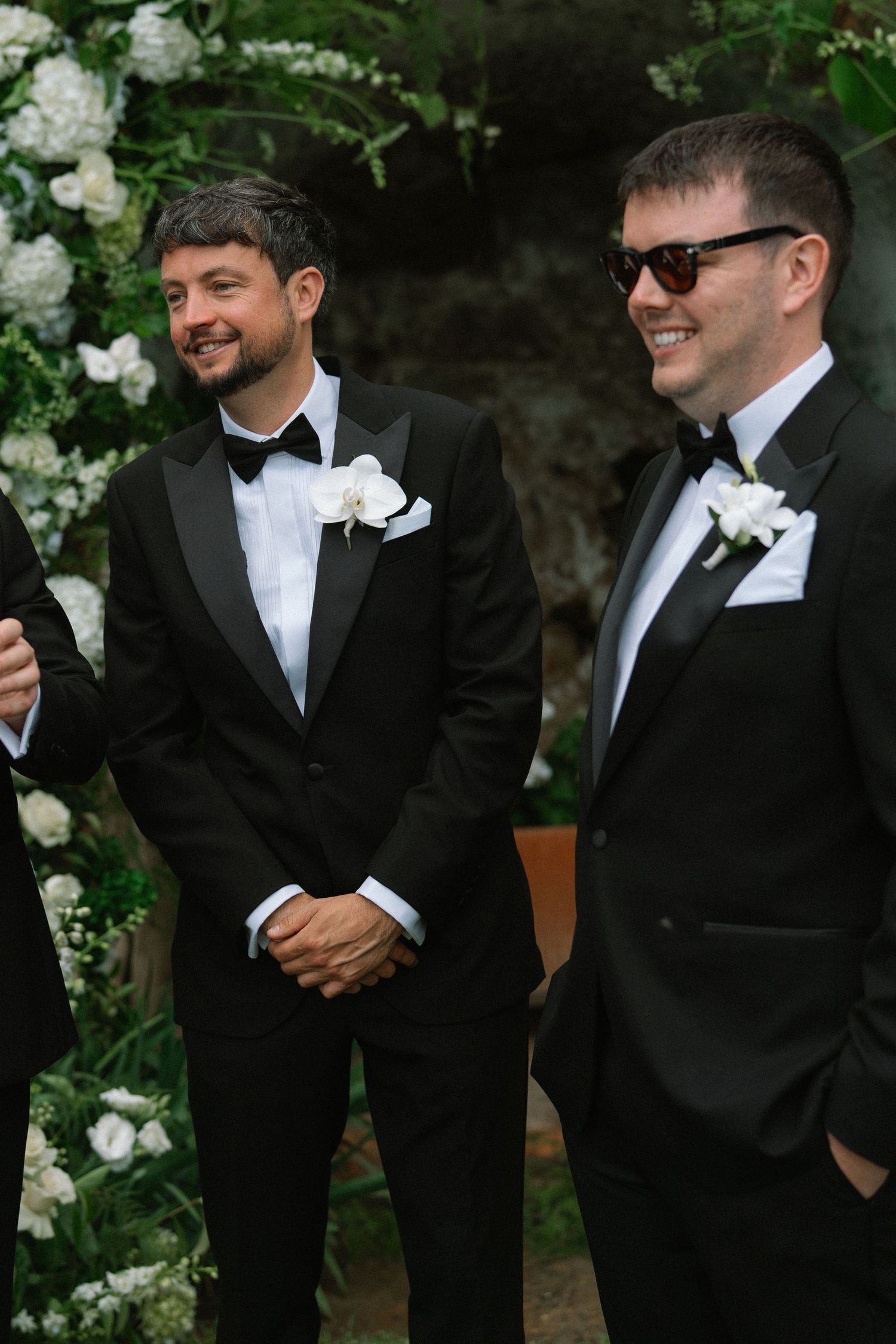 Two smiling groomsmen in black tuxedos with white boutonnieres standing at an outdoor wedding ceremony altar.