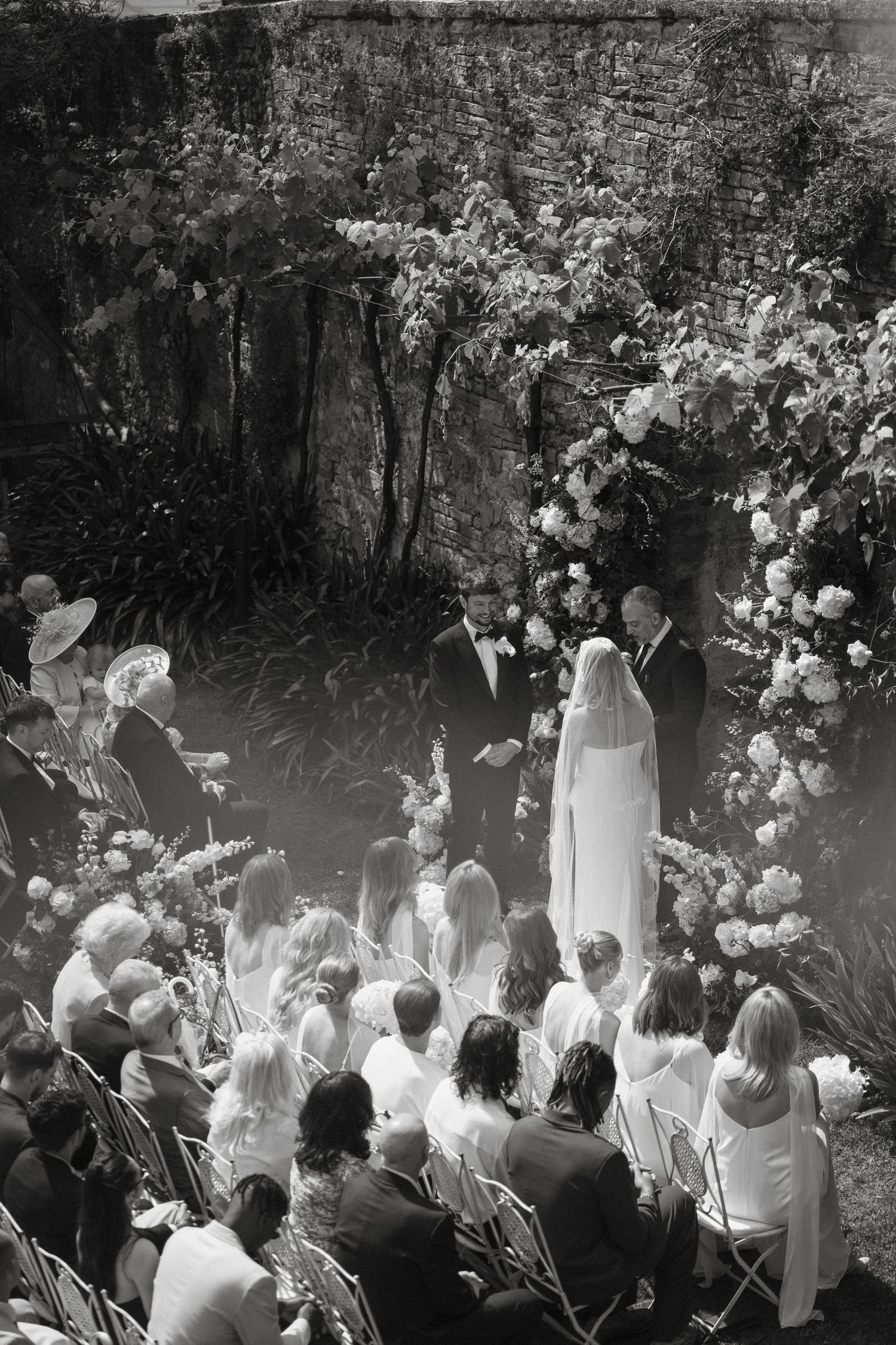Black and white aerial photo of outdoor wedding ceremony with bride and groom at floral arch before seated guests.