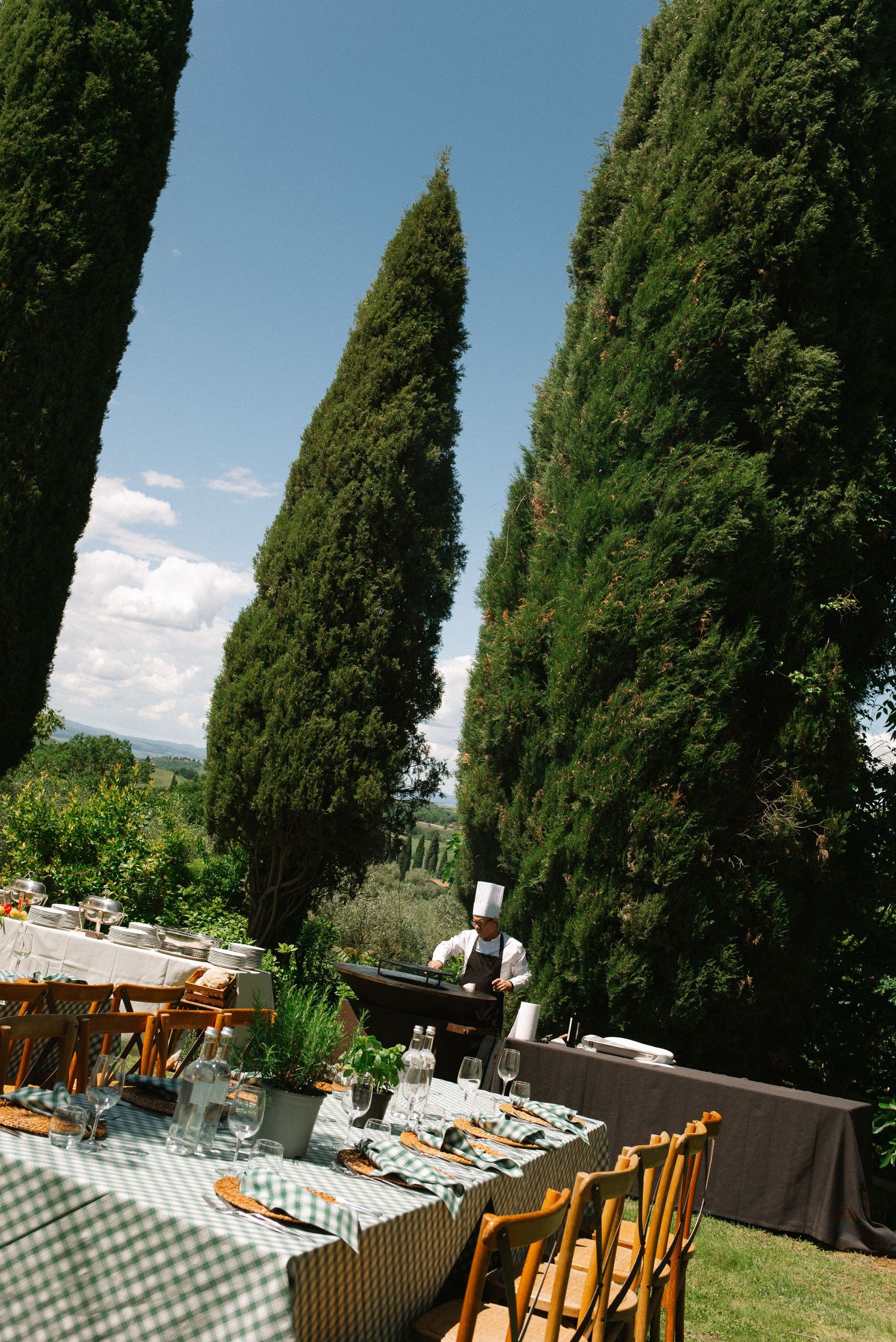 Elegant outdoor wedding reception table set among tall cypress trees in a sunny Tuscan landscape.