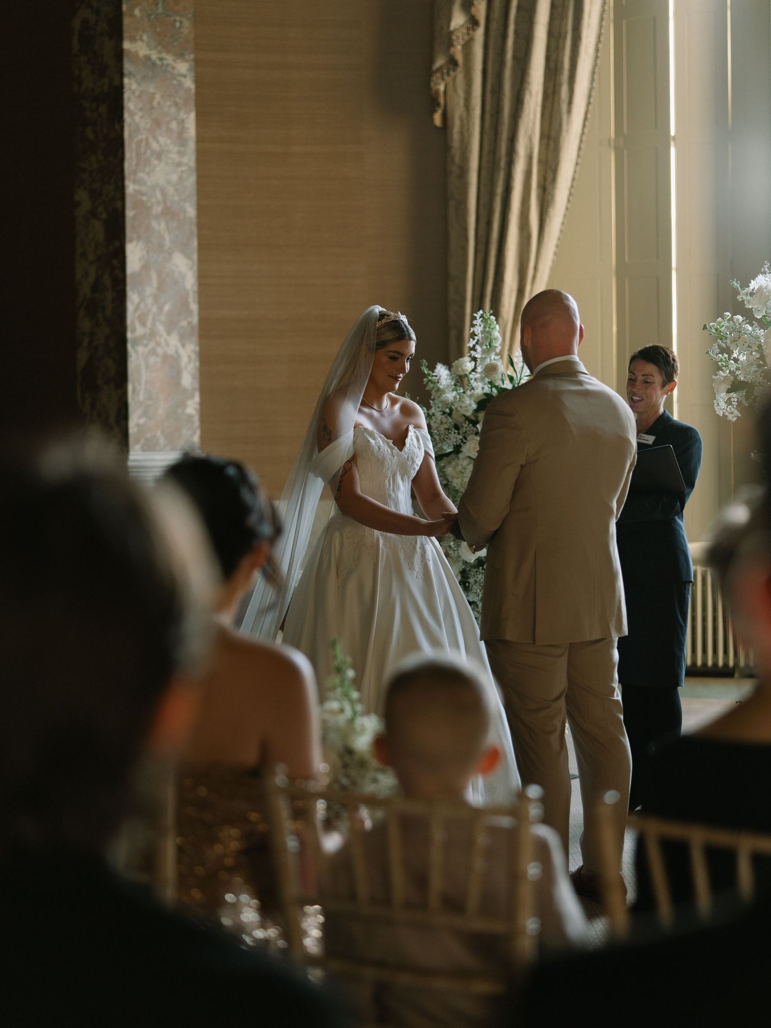 Bride in white gown and veil exchanges vows with groom at elegant indoor wedding ceremony with floral arrangements.
